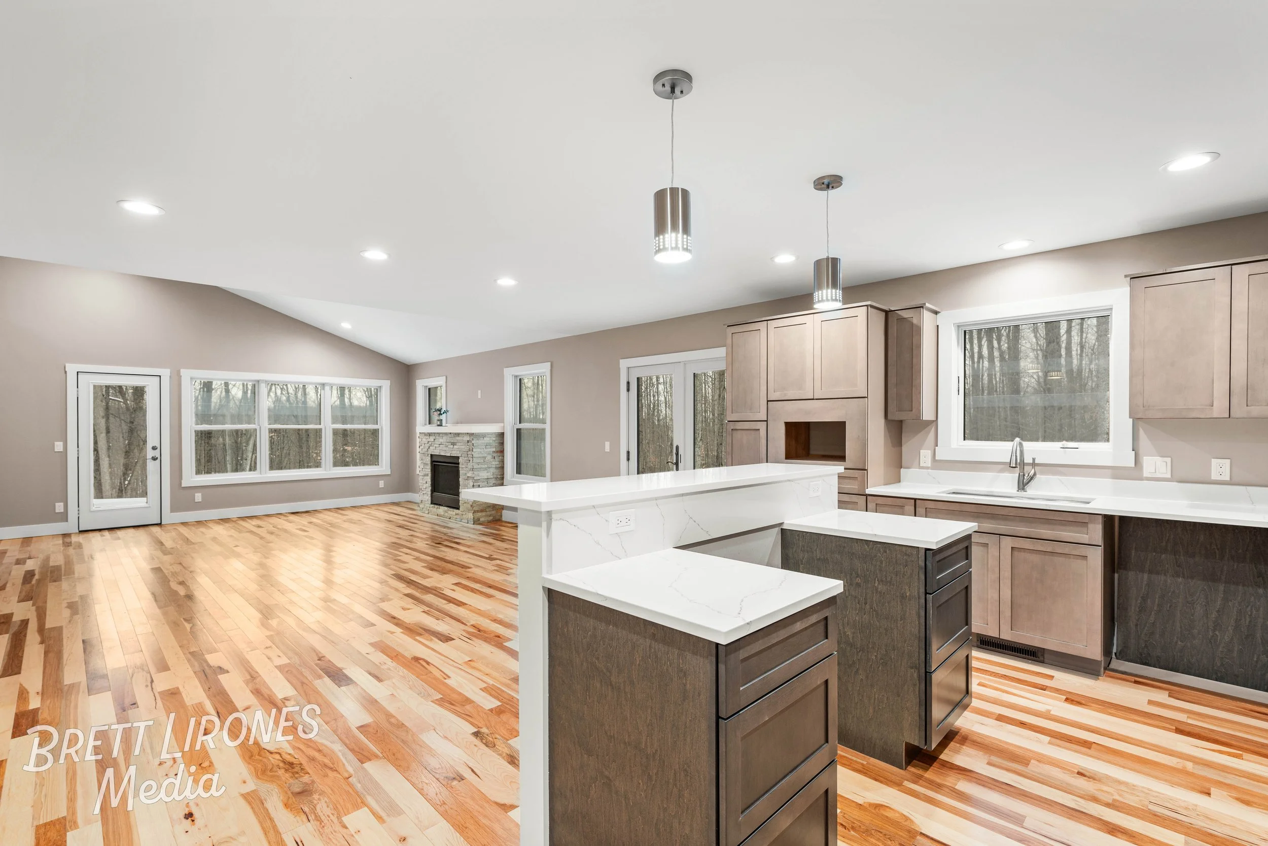Open-concept living space with hardwood floors, large windows, a stone fireplace, a kitchen with white countertops, and light wood cabinetry.