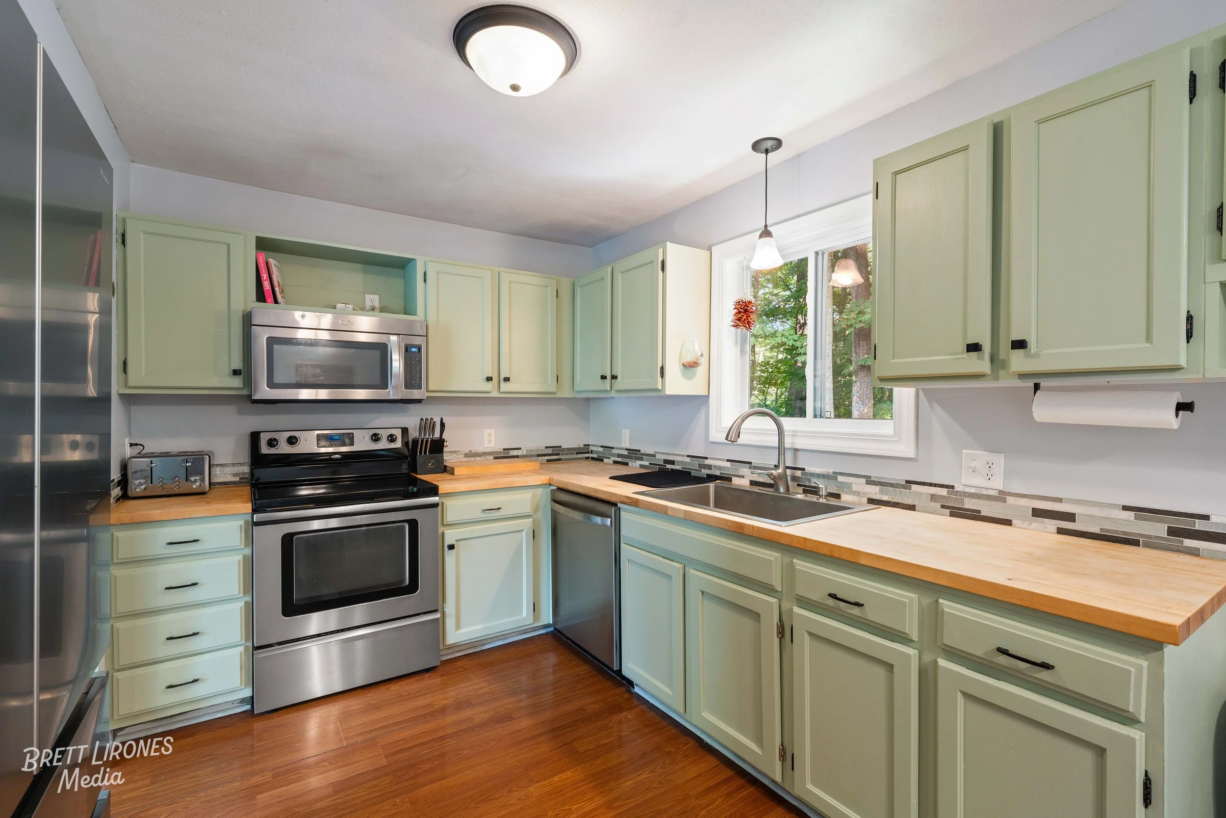 Kitchen with green cabinets, wooden countertops, stainless steel appliances including a stove and microwave, a window with trees outside, and a hardwood floor.