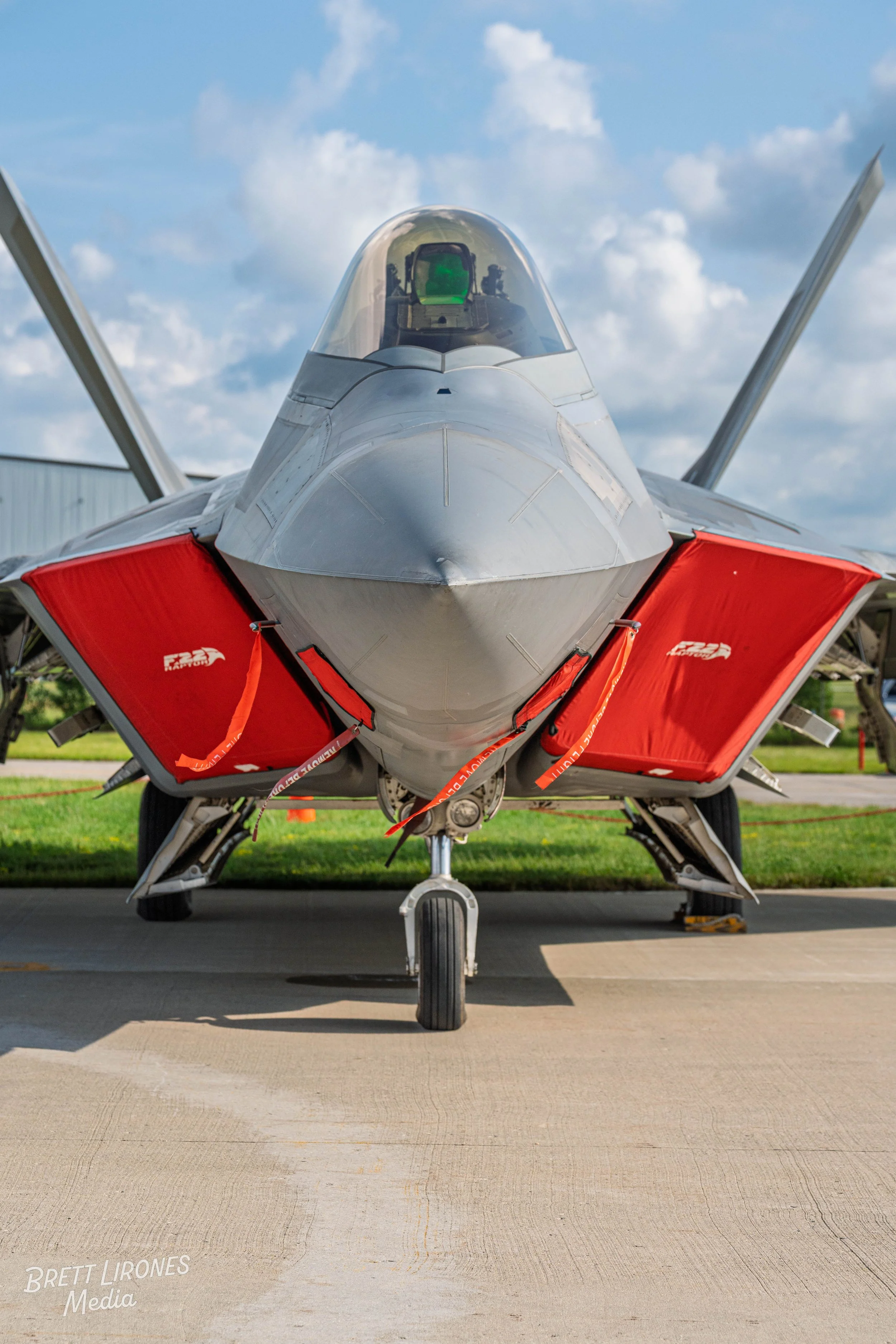 Front view of a fighter jet with red covers on its landing gear doors, parked on a tarmac under a partly cloudy sky.