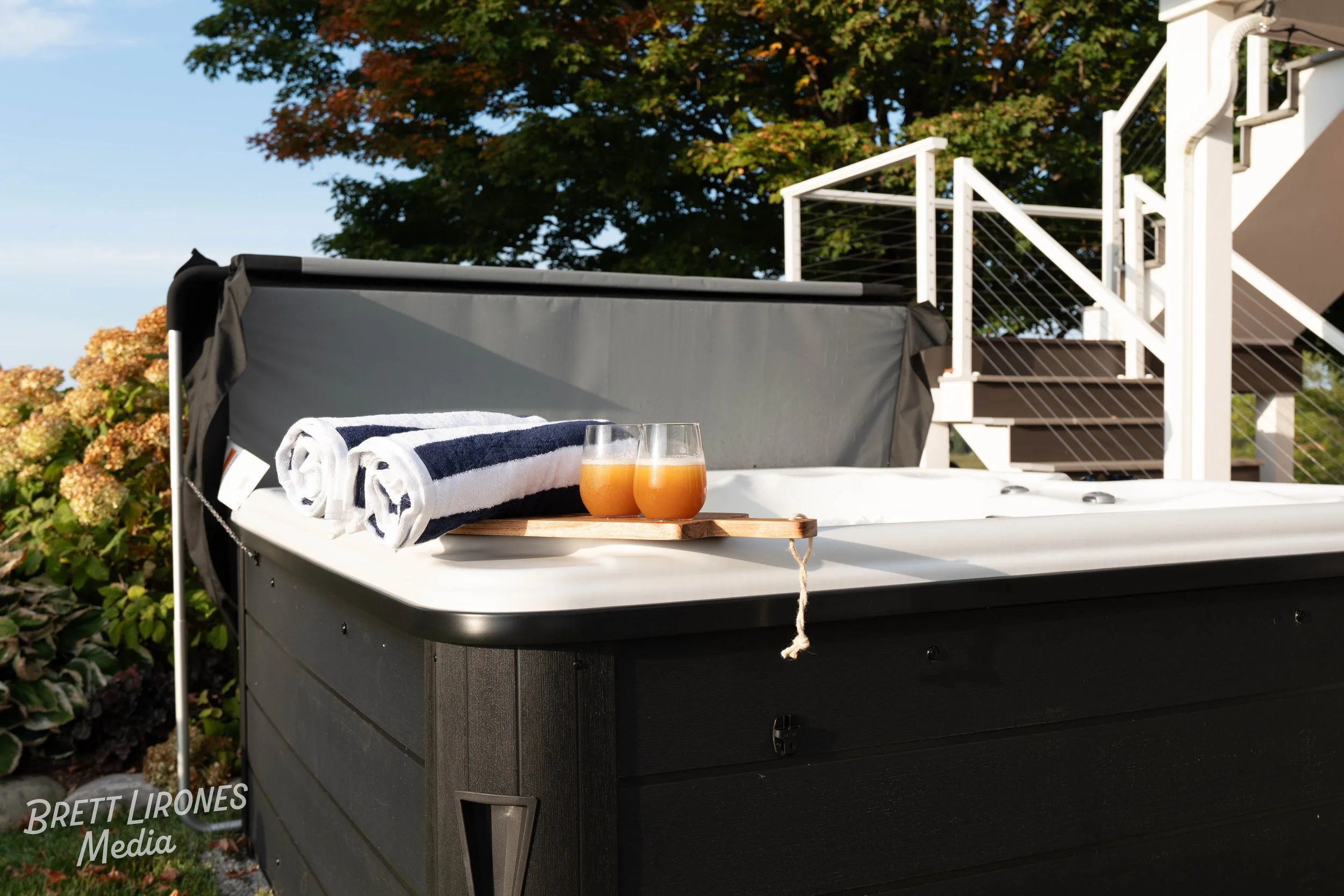 Hot tub with rolled towels and two glasses of orange juice on a wooden tray outside on a sunny day, with stairs and trees in the background.