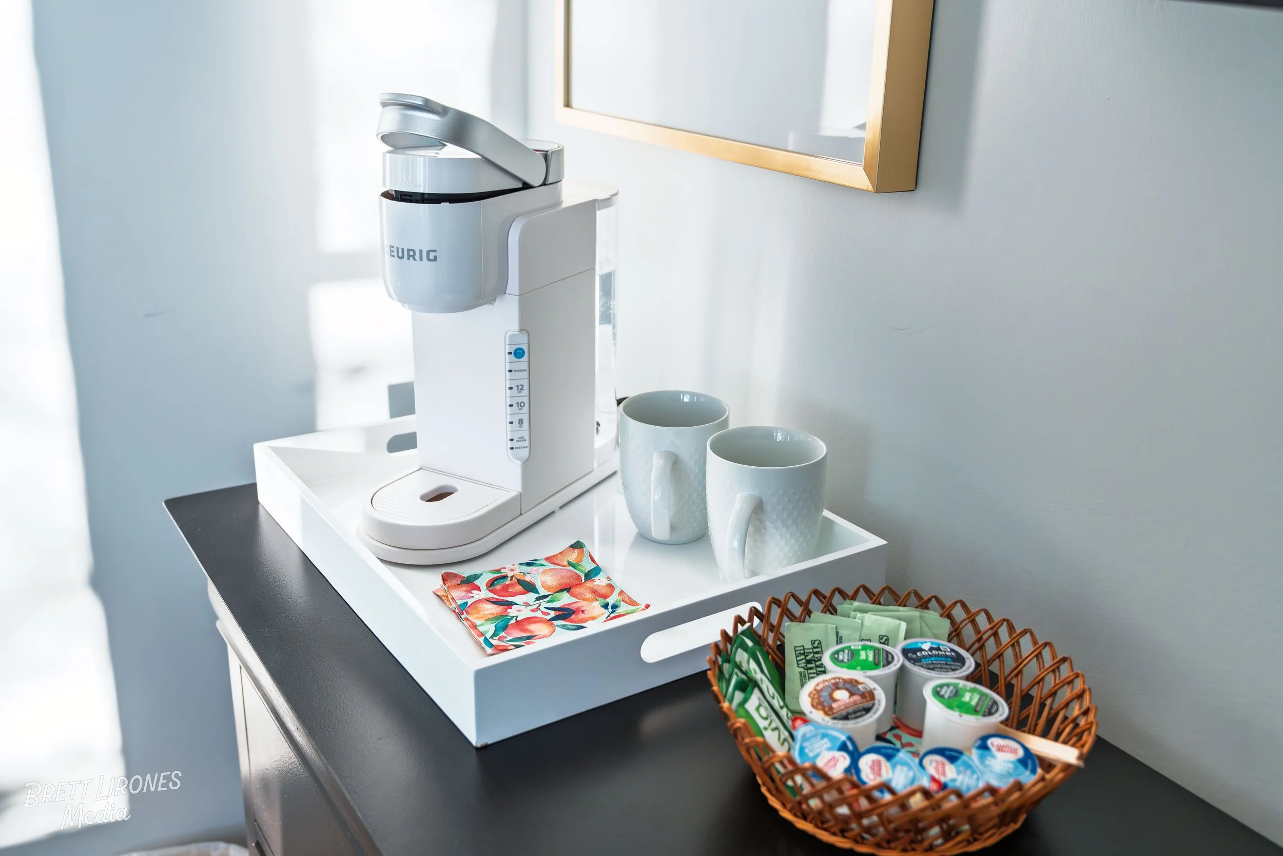 Energizer coffee machine on a black countertop with two white coffee mugs, wrapped in a white tray, a colorful napkin, and a basket with assorted small coffee condiments and creamer containers in a bright room with white walls and a framed mirror.