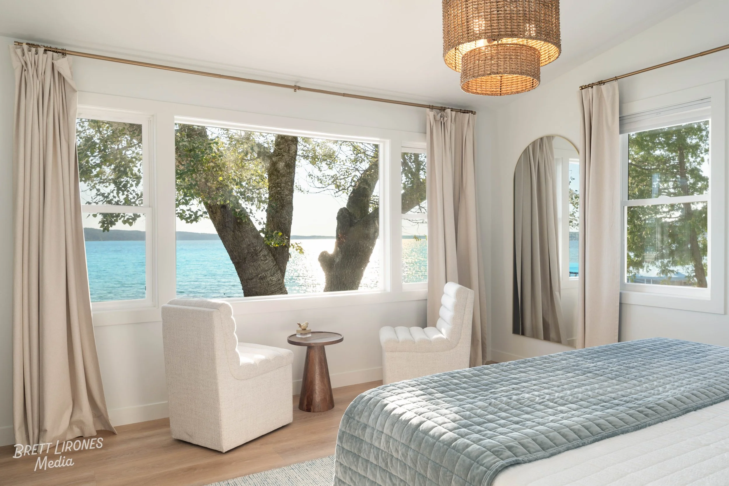 A bedroom with large windows showing a lake view and trees outside, two beige armchairs with a small round wooden table between them, and a bed with a blue quilt. A woven pendant light hangs from the ceiling, and a tall mirror is against the wall.