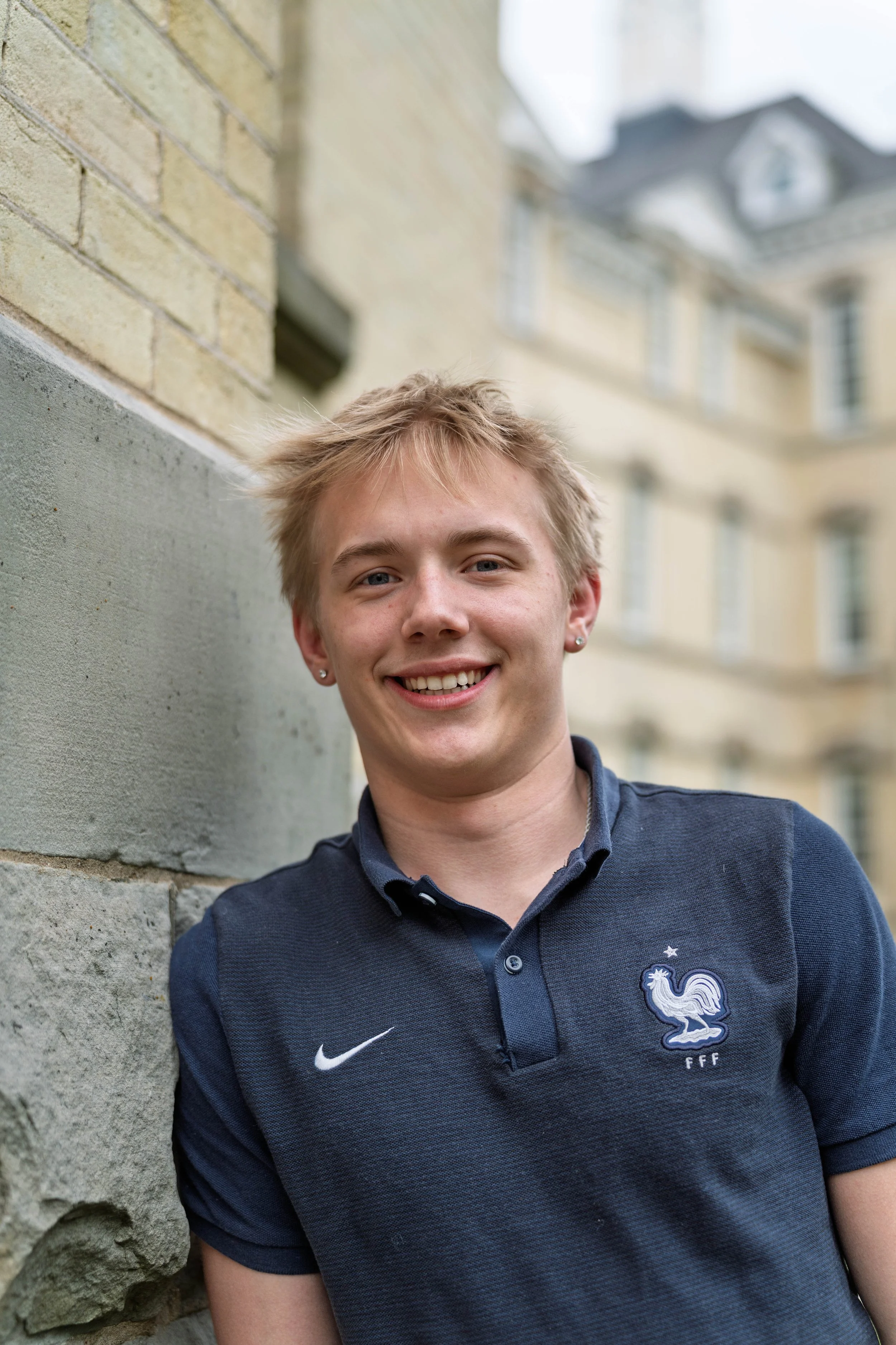 A young man with blond hair, blue eyes, and earrings, smiling and leaning against a brick and concrete wall outdoors with a building in the background.