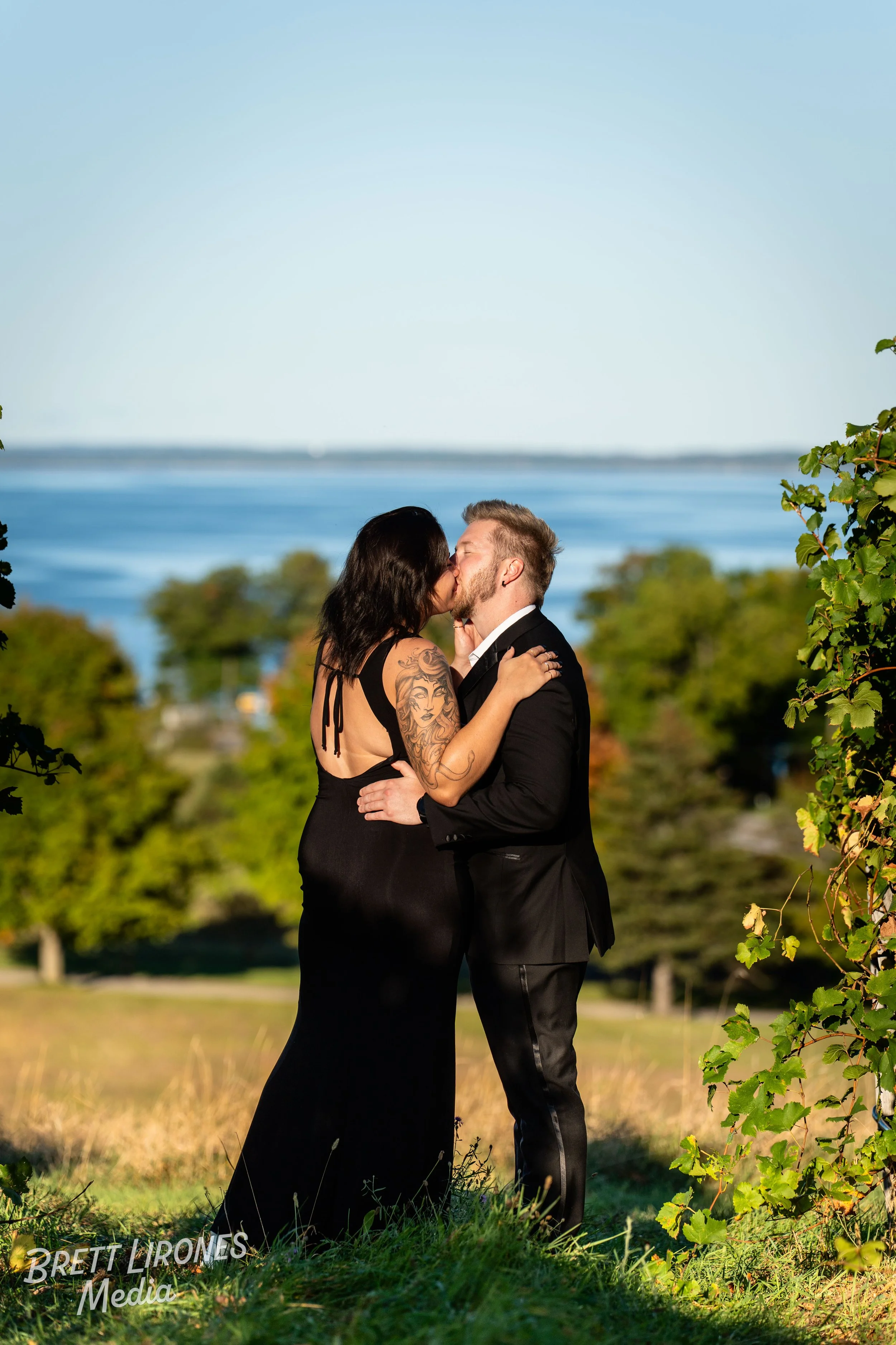 A couple in formal attire sharing a kiss outdoors, with trees and a body of water in the background.