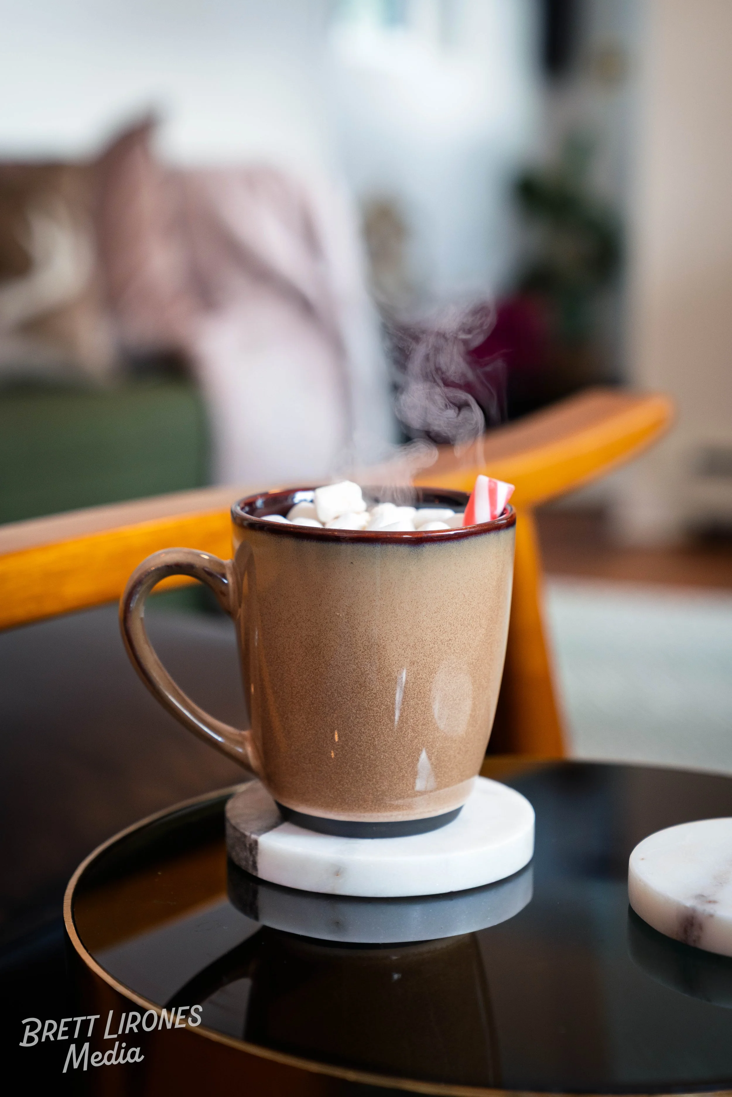 A steaming cup of hot chocolate topped with mini marshmallows and a candy cane on a black tray with a marble coaster, in a cozy indoor setting.