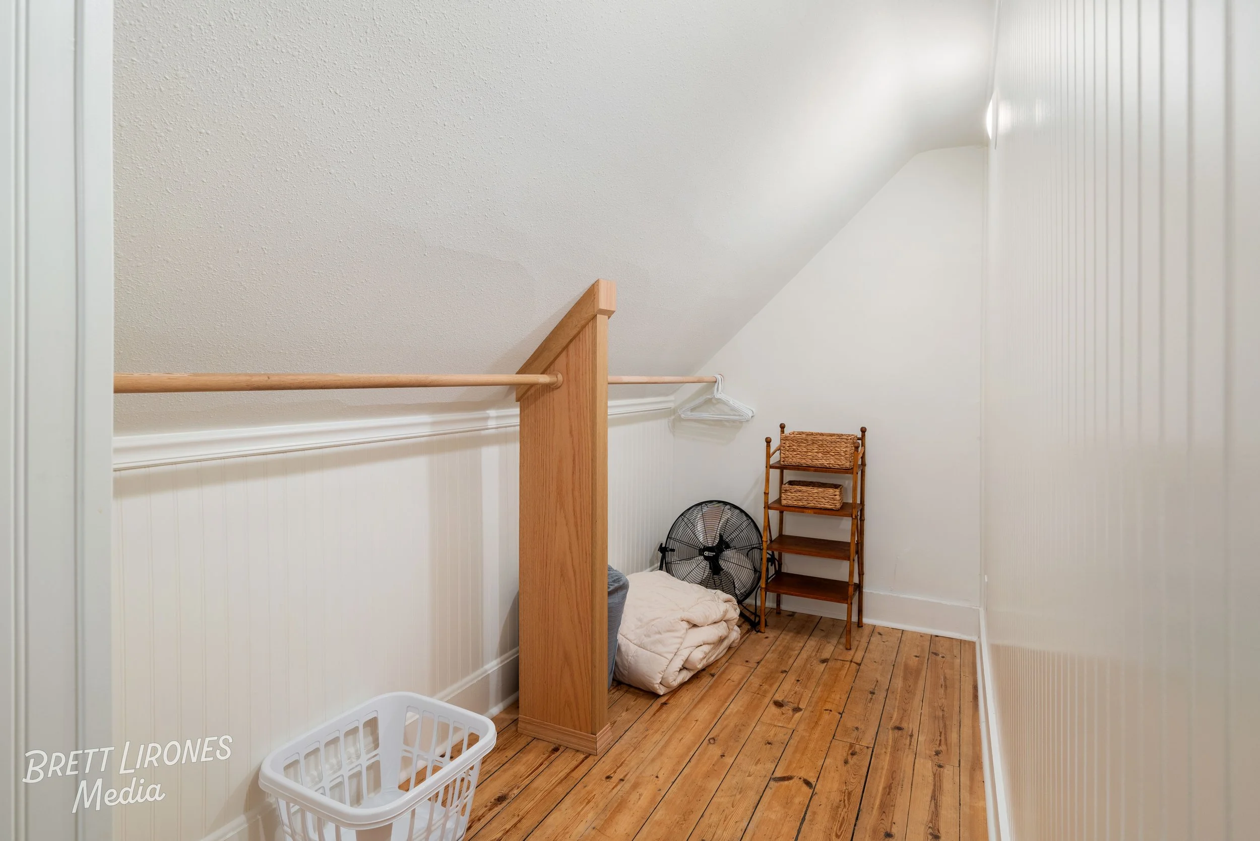 A small attic storage room with wood flooring, white paneled walls, and sloped ceiling. Contains a wicker shelf with baskets, a standing fan, a pile of blankets, a laundry basket, and some hangers.