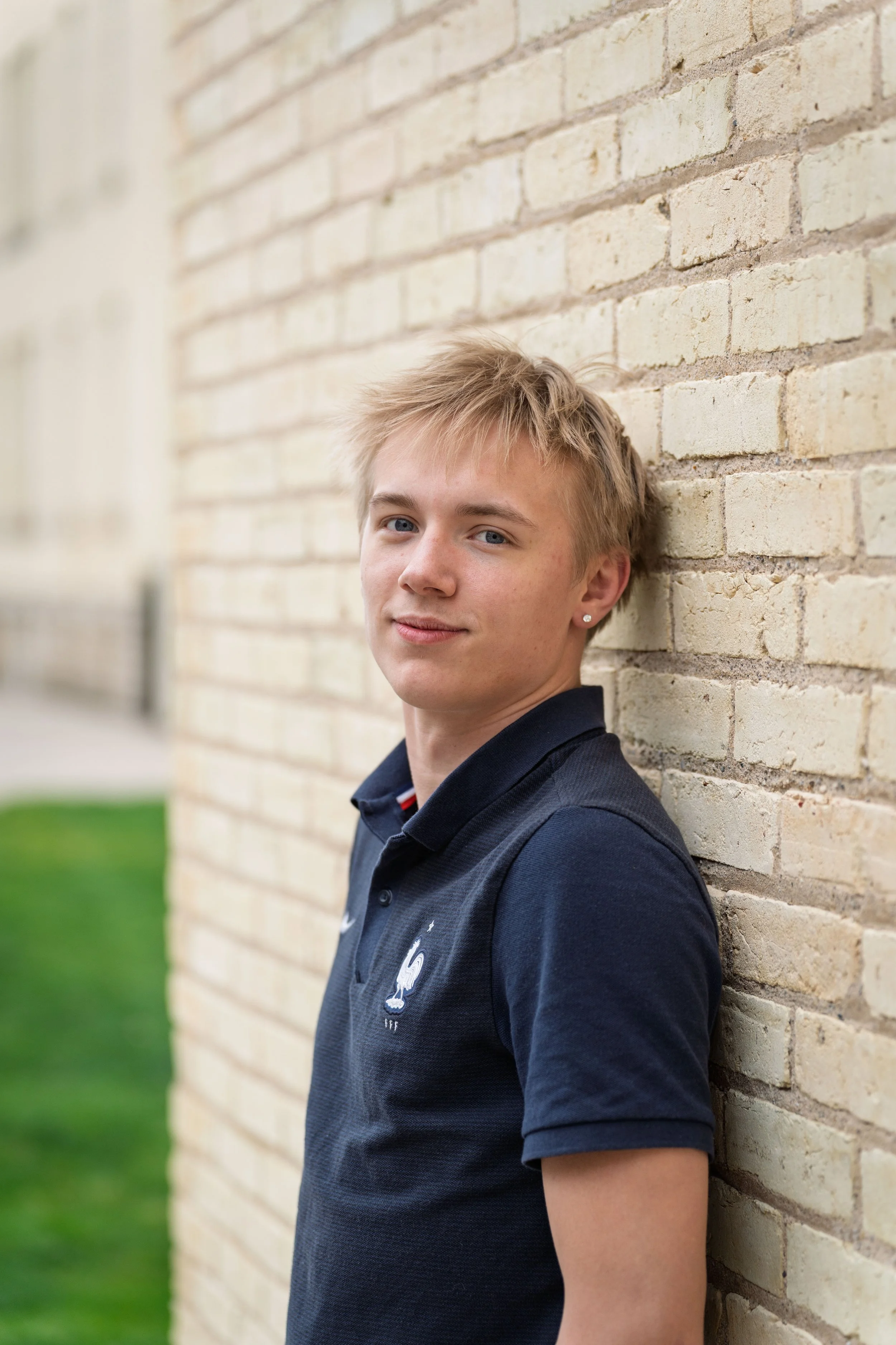 A young man with short blonde hair and earrings leaning against a beige brick wall, wearing a navy blue polo shirt with a French rooster emblem.