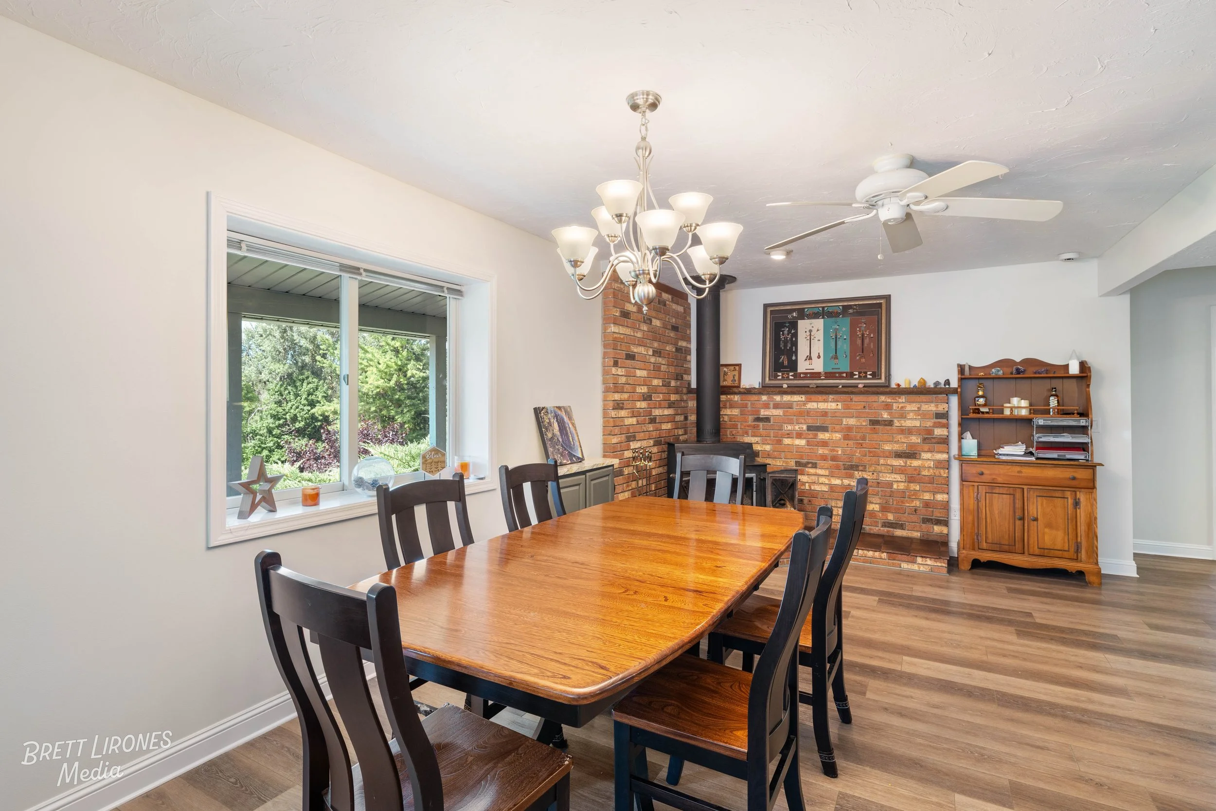 Dining room with a rectangular wooden table and six black chairs, large window with view of greenery outside, brick fireplace, wooden hutch, chandelier, and ceiling fan.