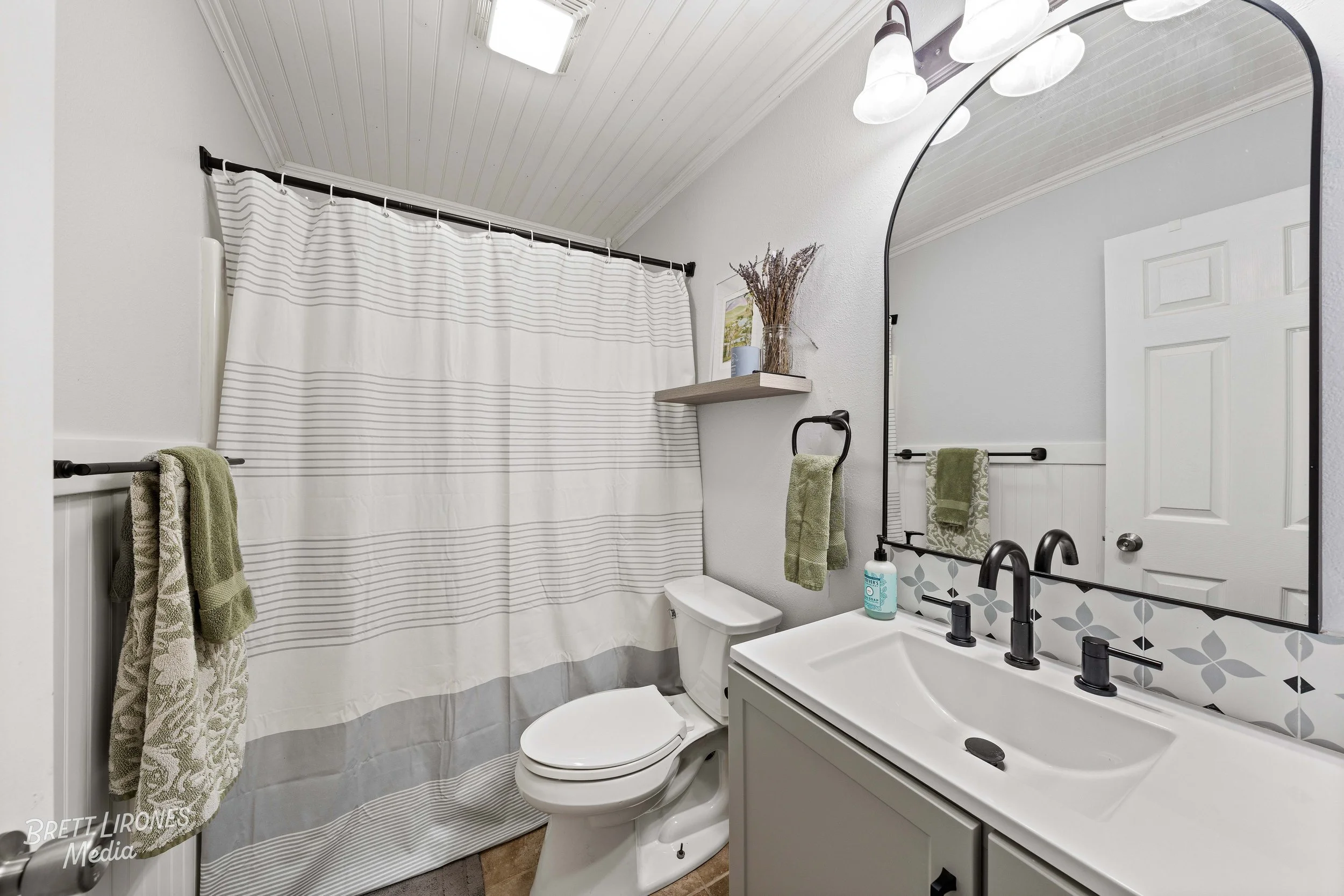 Bathroom with a white toilet, a sink with black faucet, a large mirror, a shower with a white and gray striped curtain, green towels, a wall-mounted shelf with a vase of dried flowers and a small framed picture.