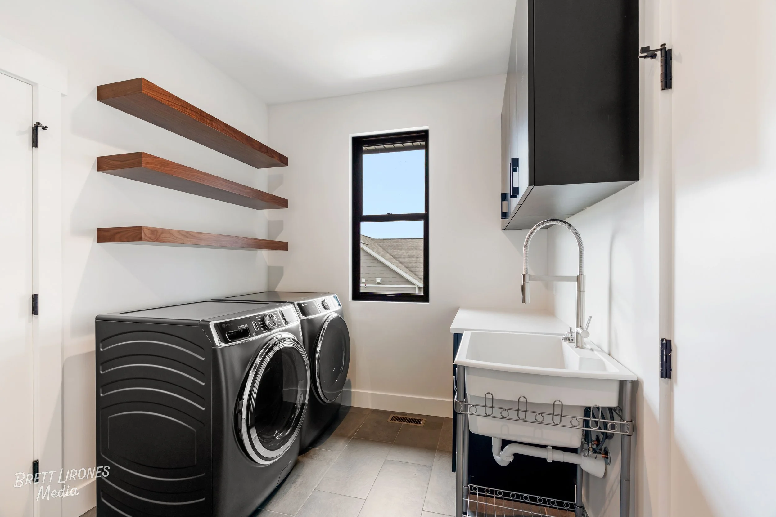 Laundry room with black washer and dryer, white utility sink, black cabinet, wood shelves, and a small window in a modern home.