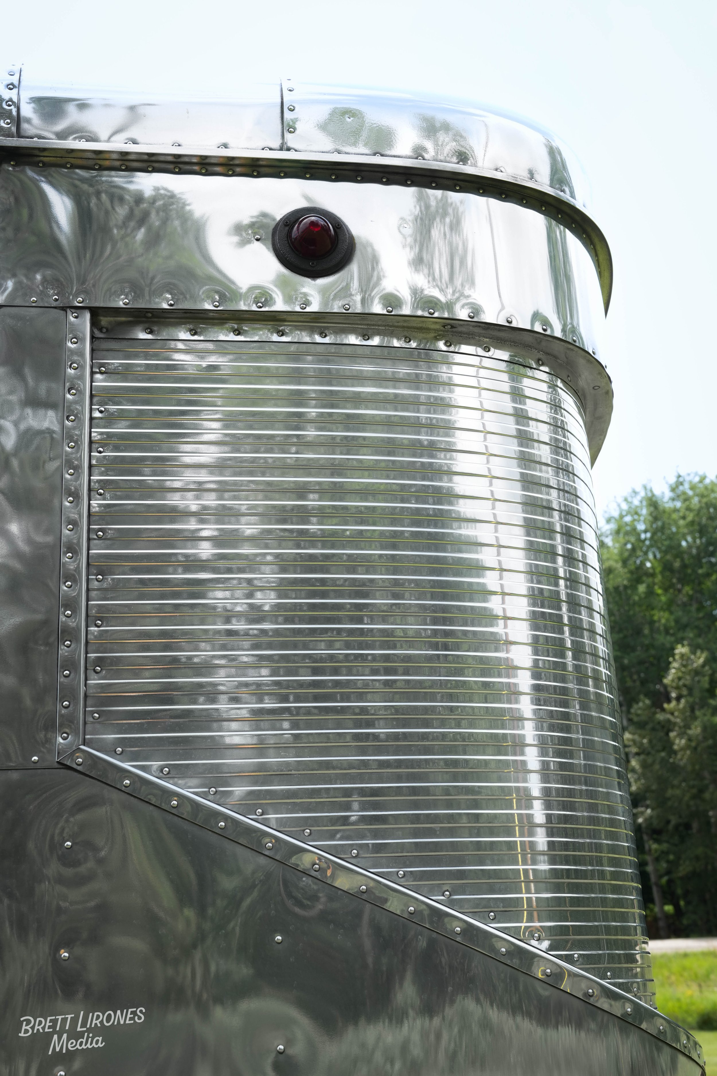 Close-up of a shiny, metallic trailer with rivets, rounded edges, and a red indicator light on the front, outdoors with trees in the background.