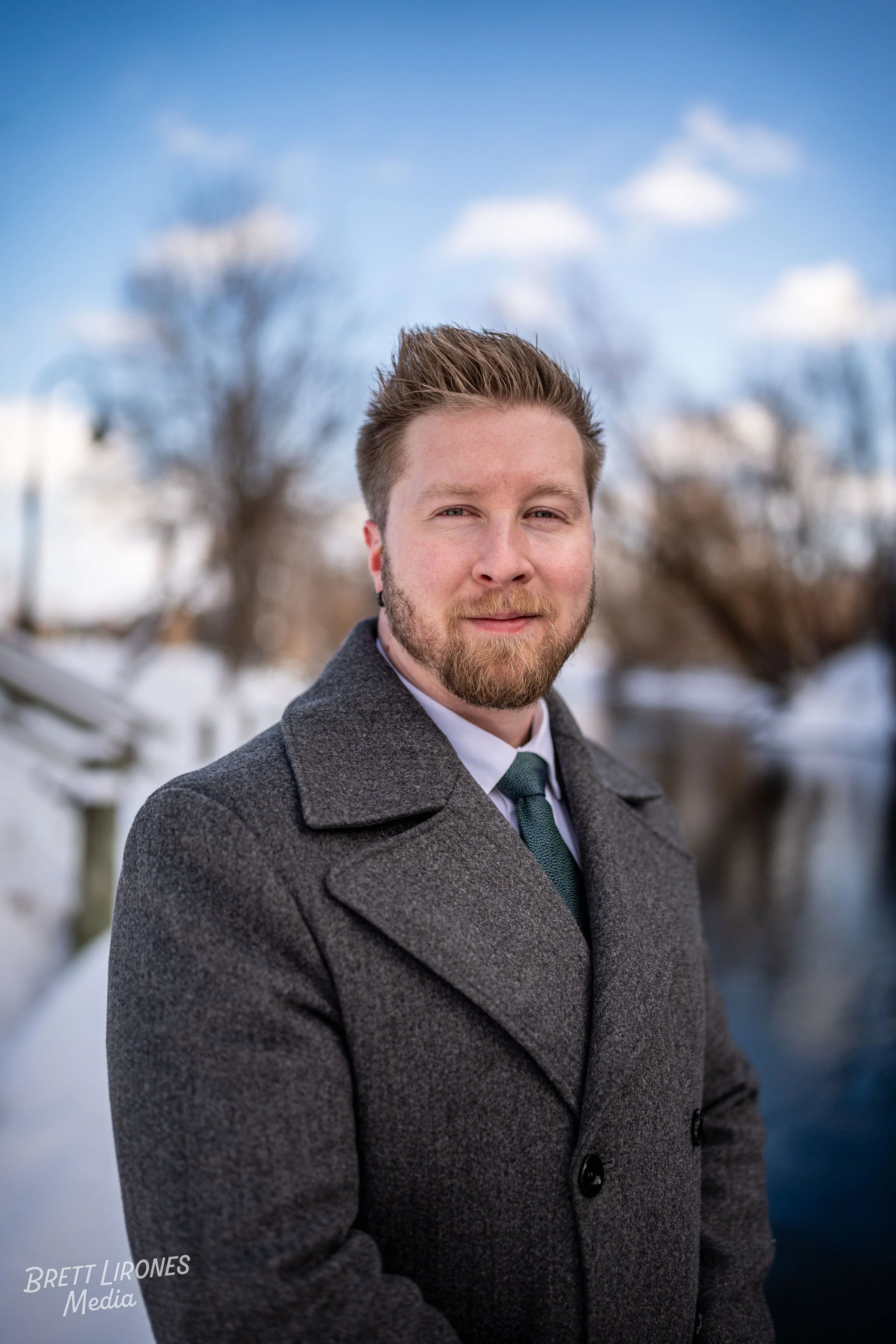 A man with light skin, a beard, and styled hair, wearing a gray coat and a dark green tie, stands outdoors near a body of water under a blue sky with scattered clouds and snow on the ground.