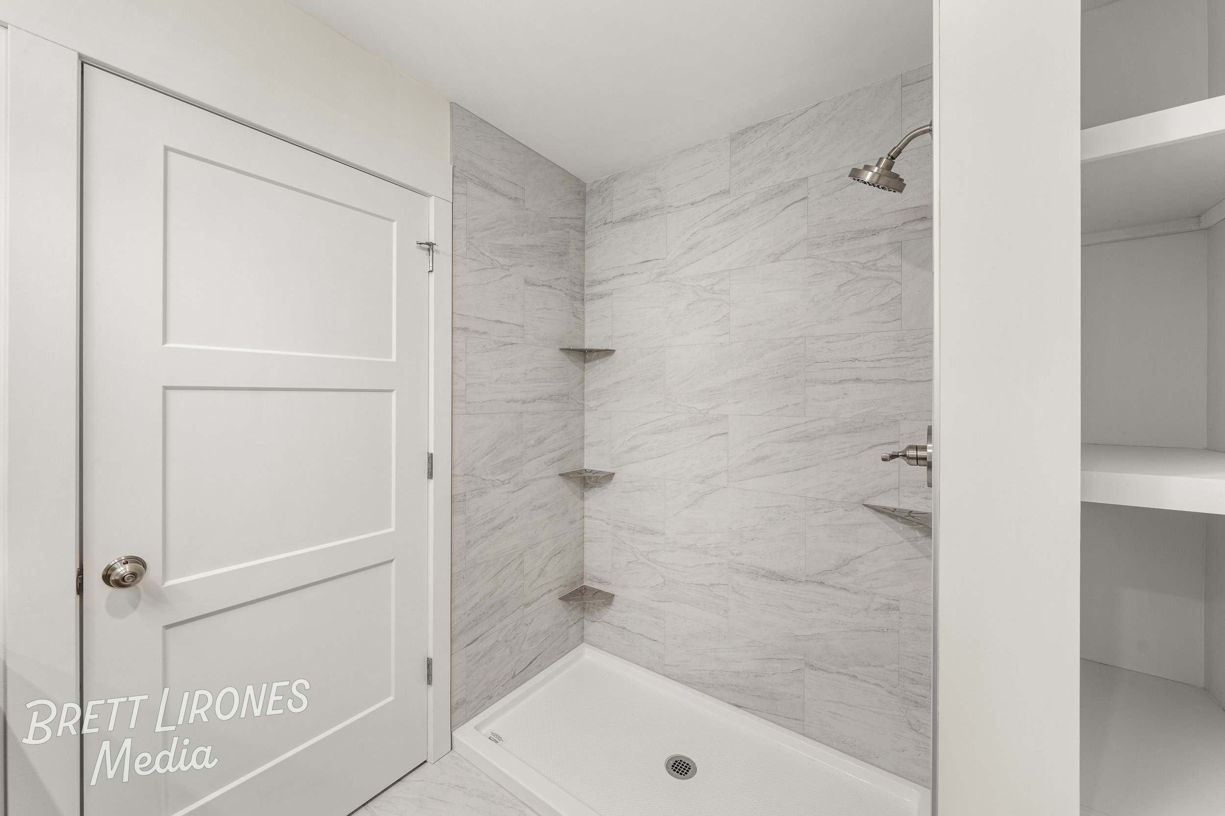Modern walk-in shower with marble tile walls, a white shower base, a round showerhead, and built-in shelves, adjacent to a white door and a white storage closet.