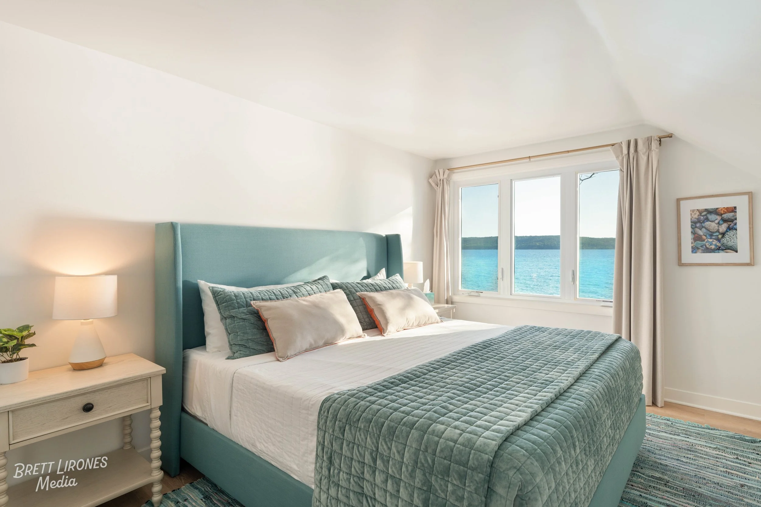 Cozy bedroom with a king-sized bed, turquoise headboard, and textured bedding, overlooking a bright window with a view of water and distant shoreline.