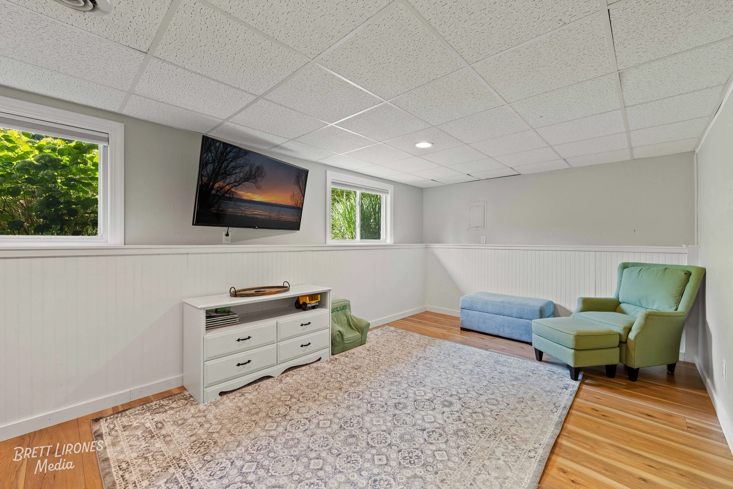 A cozy basement room with white walls, a white crocheted curtain, a wall-mounted flat-screen TV showing a sunset, two small windows with greenery view, an area rug, a green armchair with matching ottoman, a blue cushioned bench, a white dresser with 