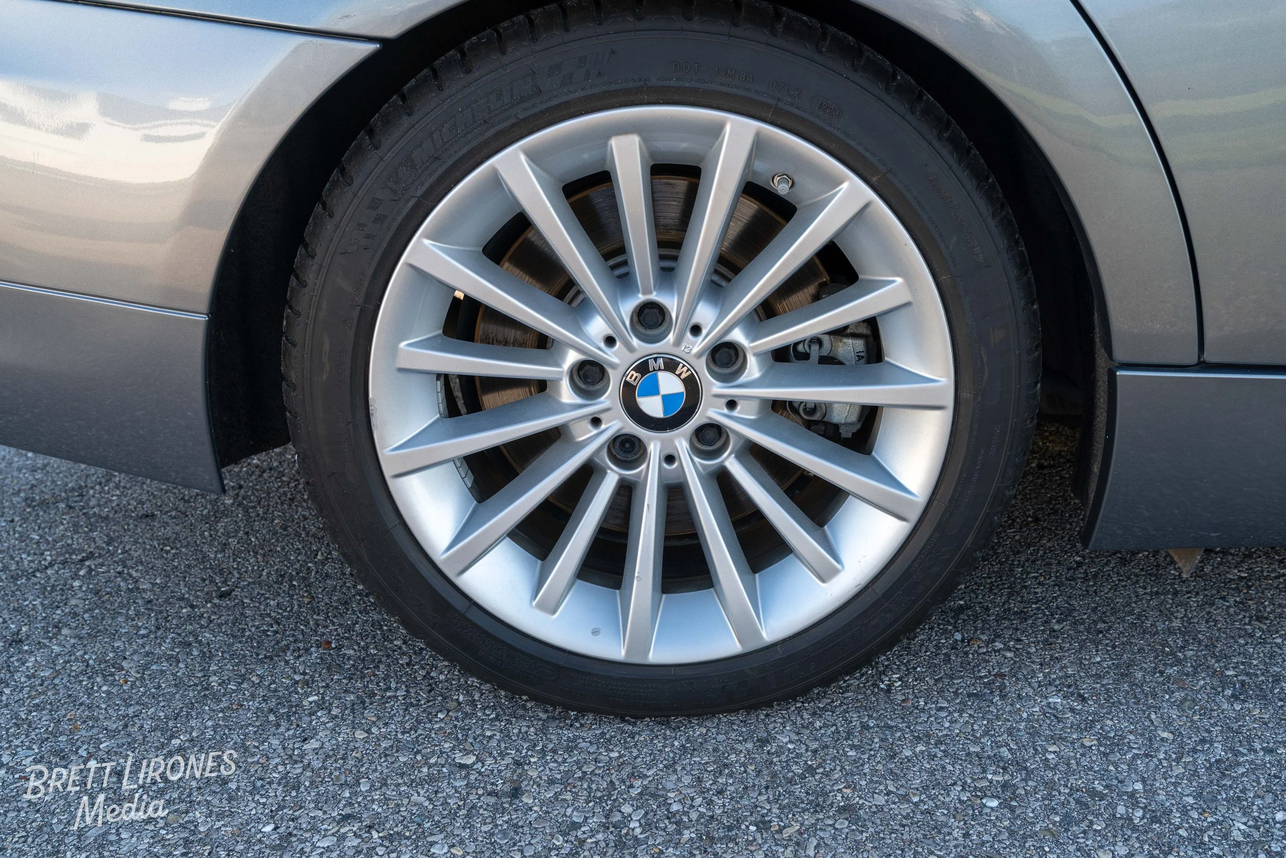Close-up of a silver BMW car wheel with a tire, showing the BMW logo in the center of the alloy rim, parked on a gravel surface.