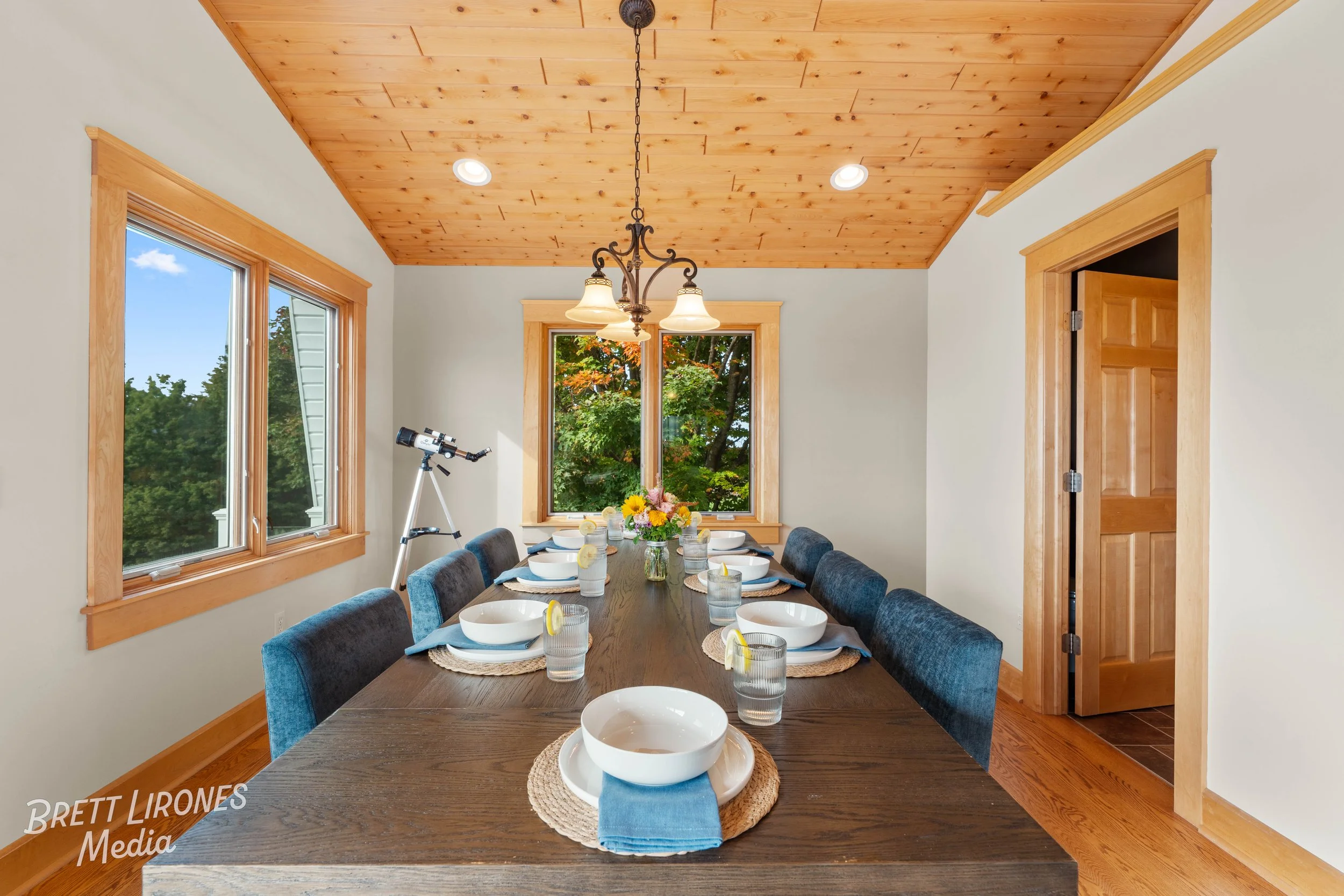A dining room with a wooden ceiling, large window showing trees outside, and a long wooden table set with bowls, glasses with lemon slices, blue napkins, and a flower centerpiece. There's a telescope by the window and a chandelier hanging from the ce