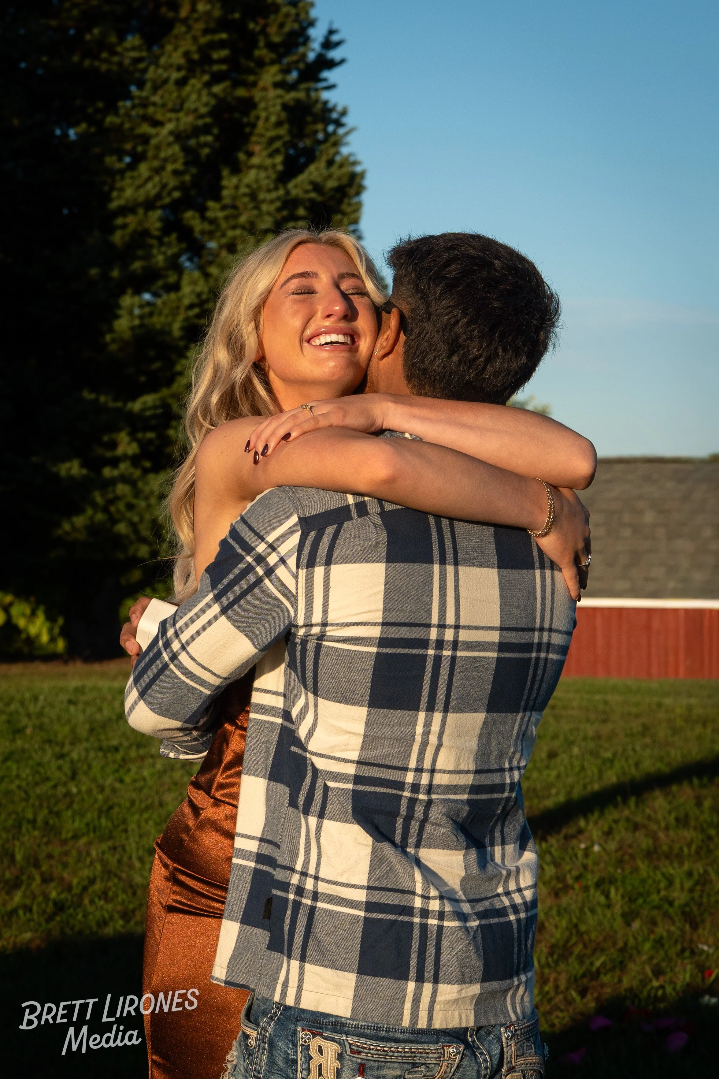 A man and woman hugging outdoors in the sunlight. The woman is smiling and has her eyes closed, wrapped around the man's neck. The man has his back to the camera and is holding her. There are trees and a building in the background.