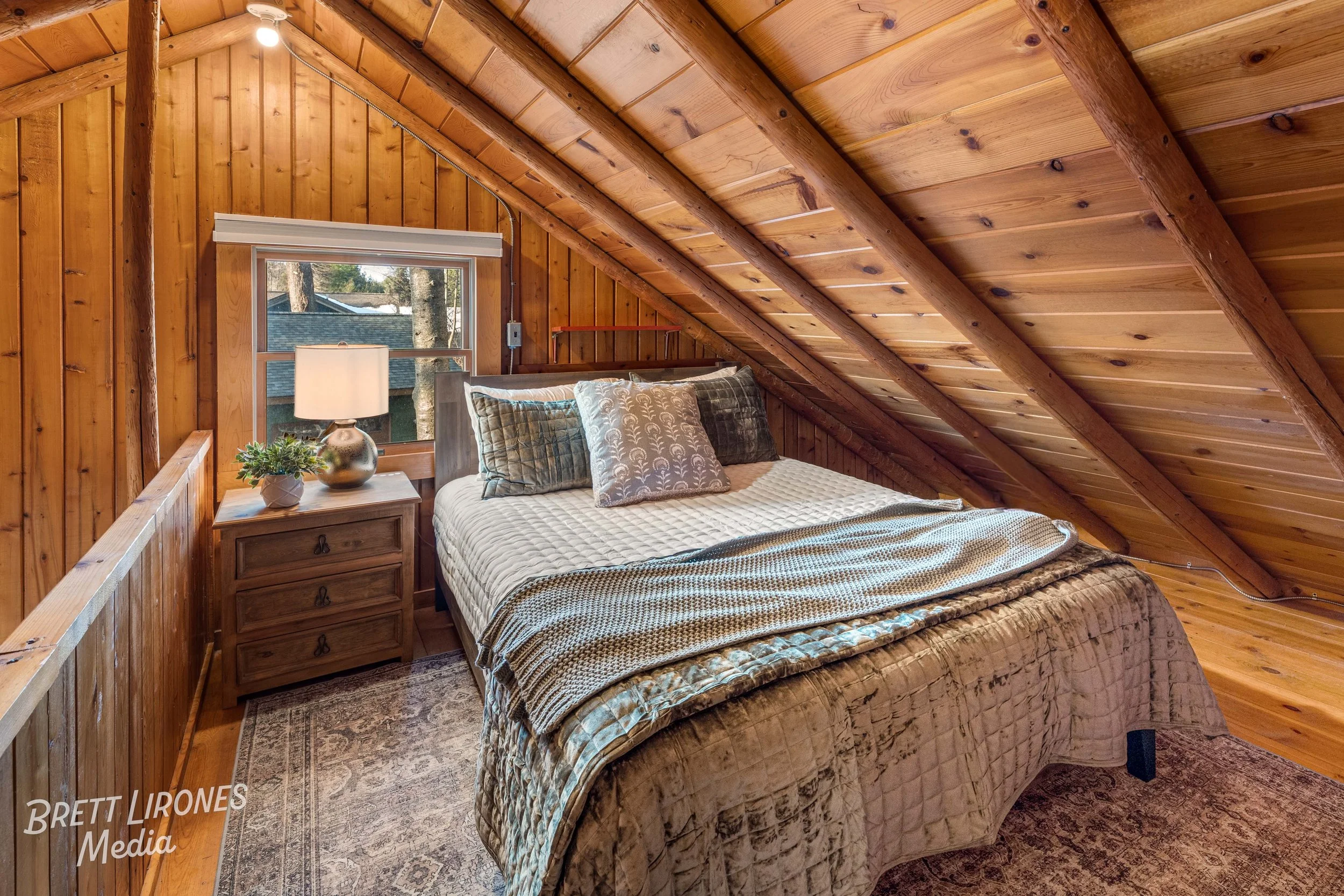 A cozy bedroom with wood-paneled walls and ceiling, a bed with multiple pillows, a nightstand with a lamp and potted plant, and a small window showing trees outside.