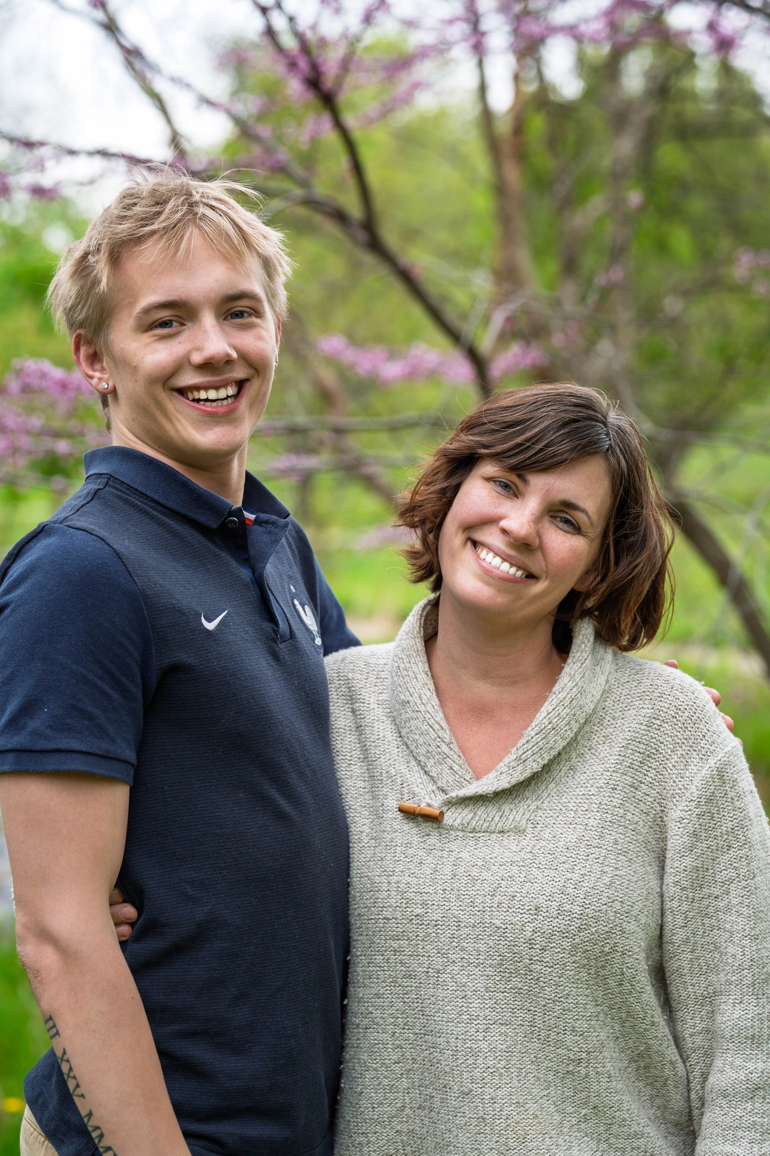 A young man and a woman smiling outdoors in a garden with blooming pink and purple flowers on trees.