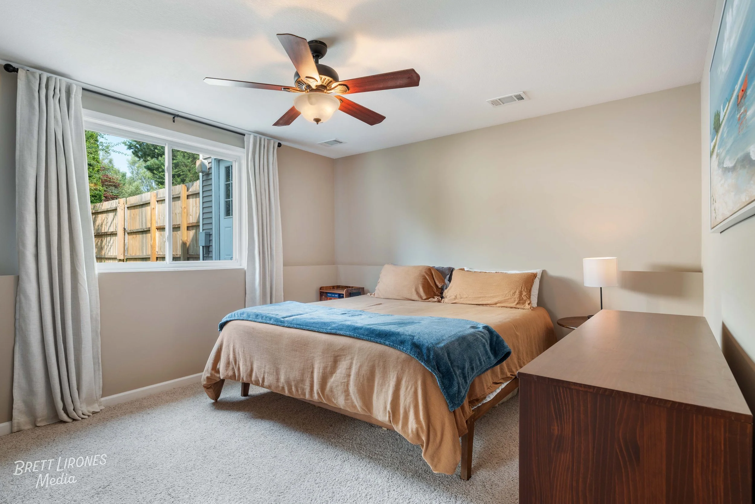 Bedroom with a bed, beige bedding, blue throw blanket, white curtains, window showing outside greenery, ceiling fan with light, wooden dresser, small nightstand with lamp, wall art, light-colored carpet floor.