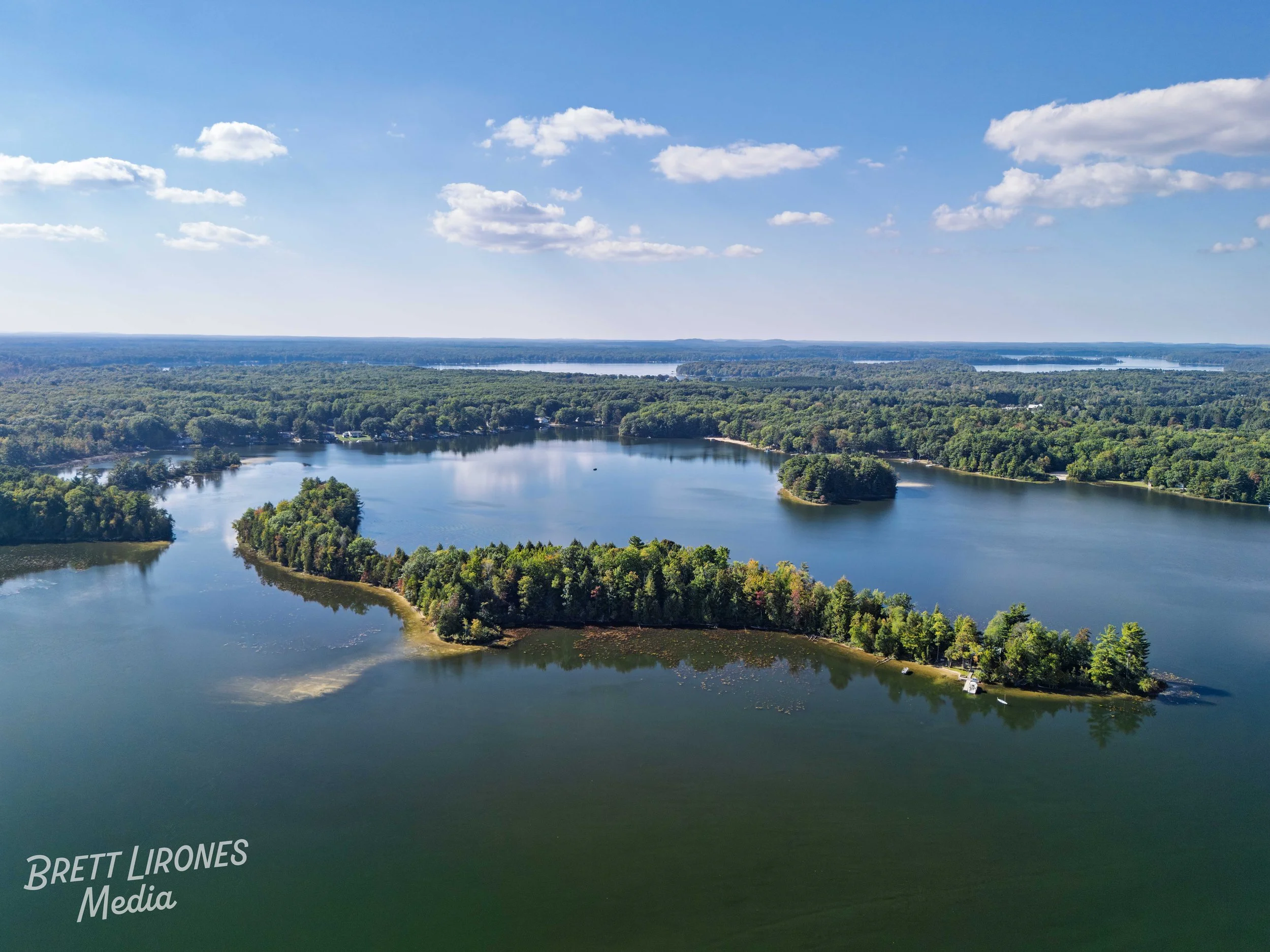 Aerial view of a large lake with multiple islands covered in green trees, surrounded by dense forest under a partly cloudy sky.