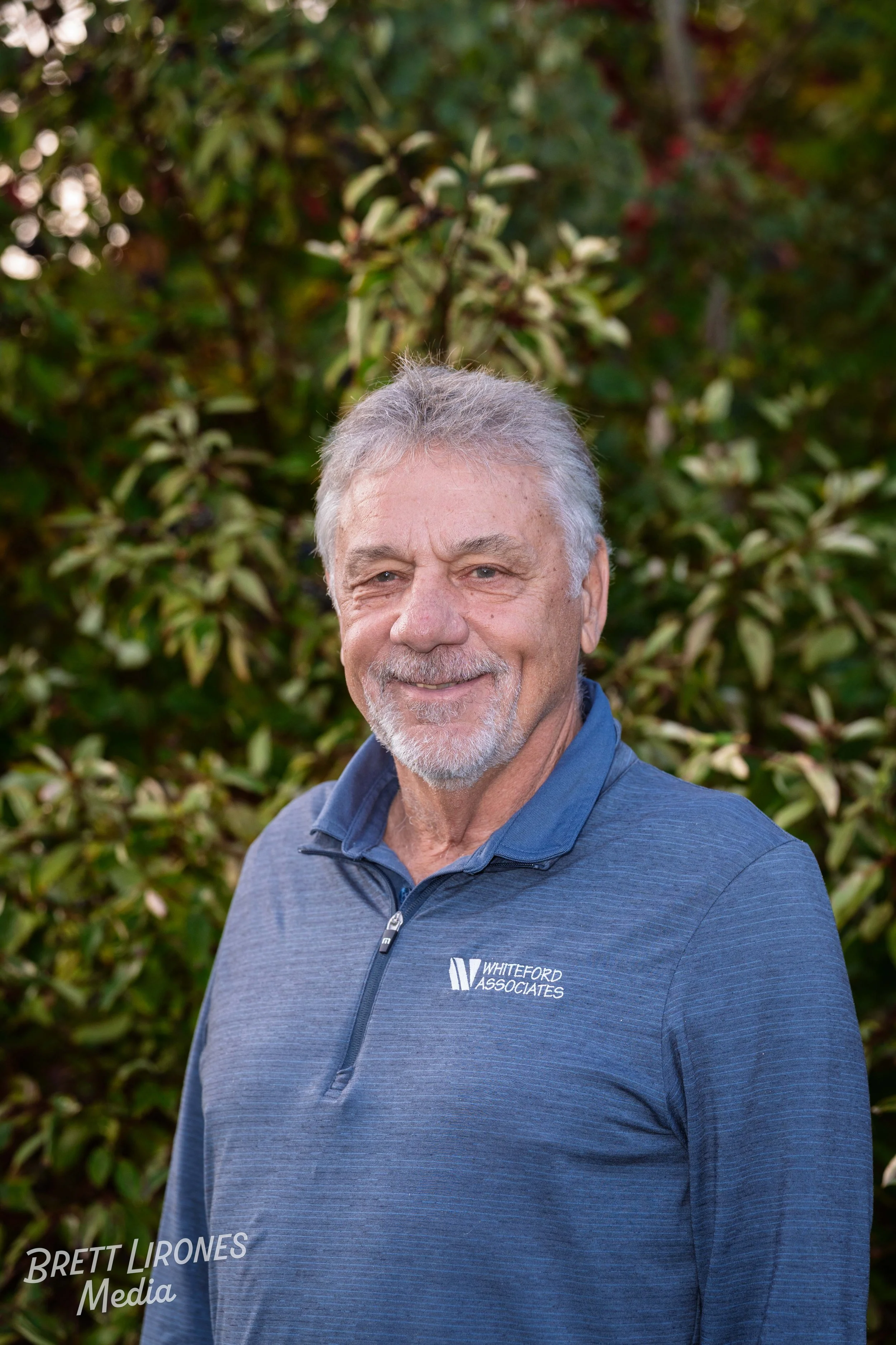 A smiling older man with gray hair and beard wearing a blue zip-up jacket standing in front of foliage.