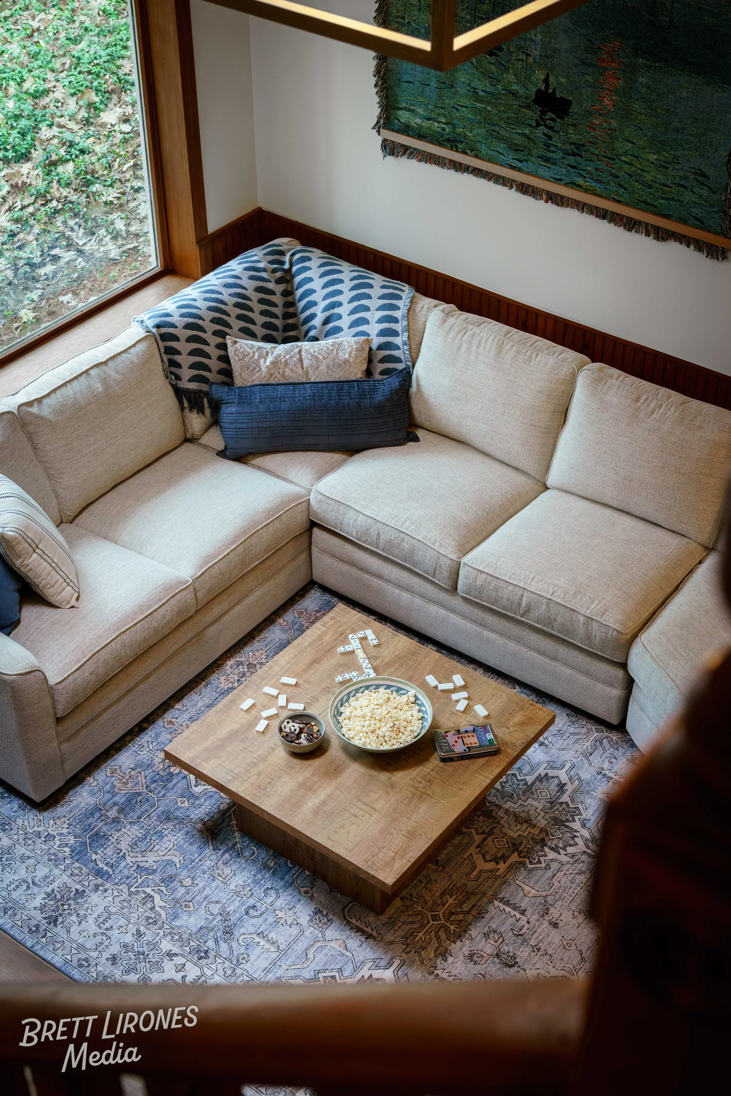A beige sectional sofa with a patterned blanket and pillows in a living room. A wooden coffee table with bowls of popcorn, snacks, and a smartphone on it. A patterned blue rug is underneath. A large window with greenery outside and a wall with a text
