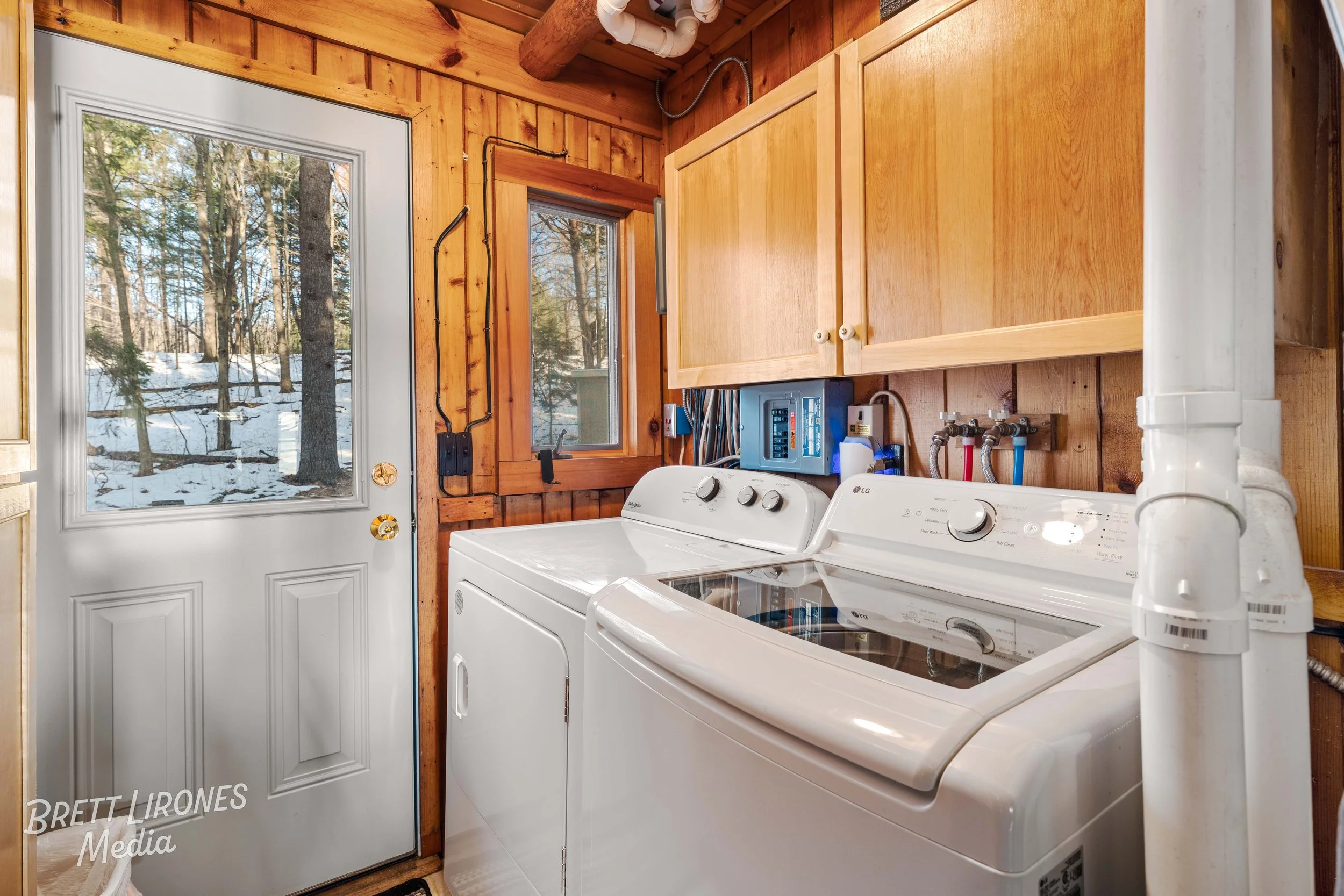 Laundry room with a washer and dryer, wooden walls, window, and door showing snowy woods outside.