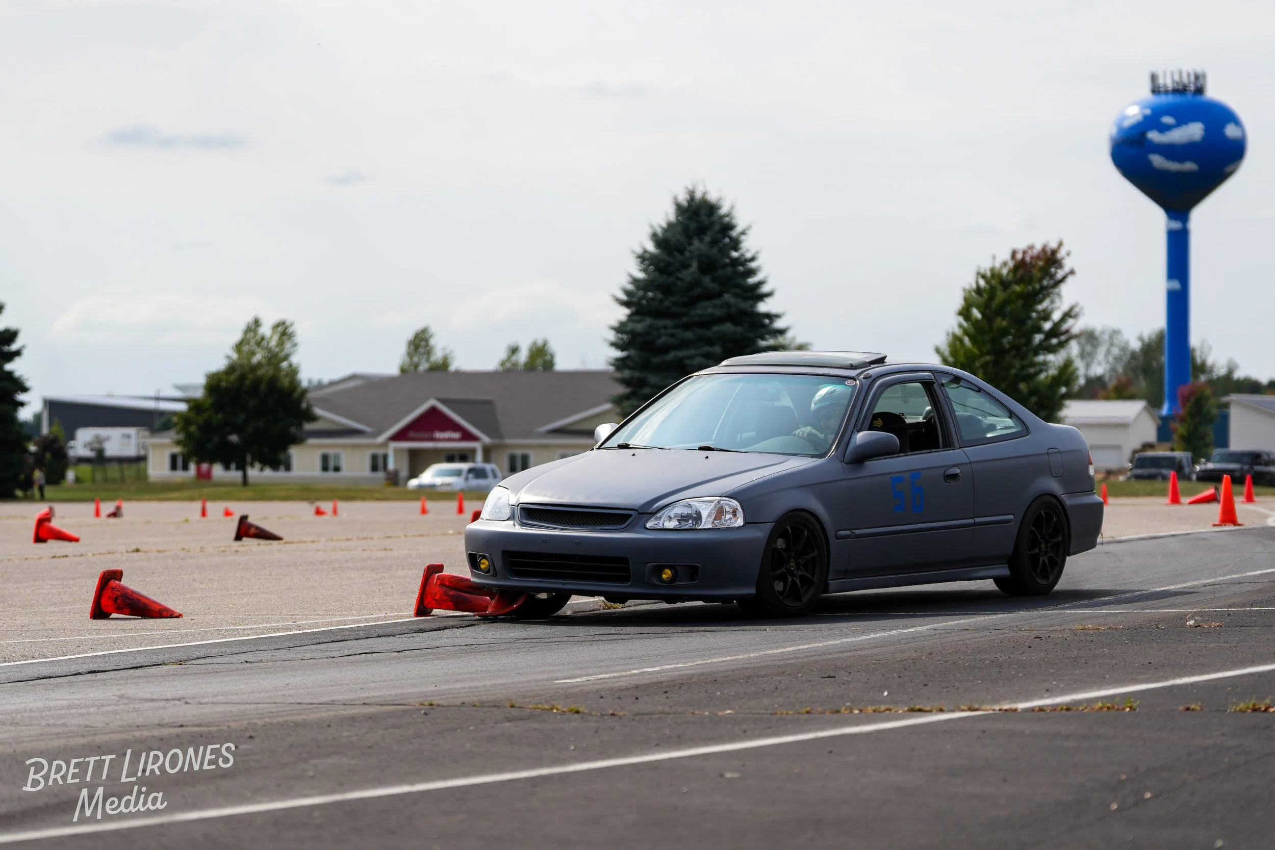A gray racing car moving through a slalom course marked by orange cones on an open parking lot or airstrip with a water tower and buildings in the background.