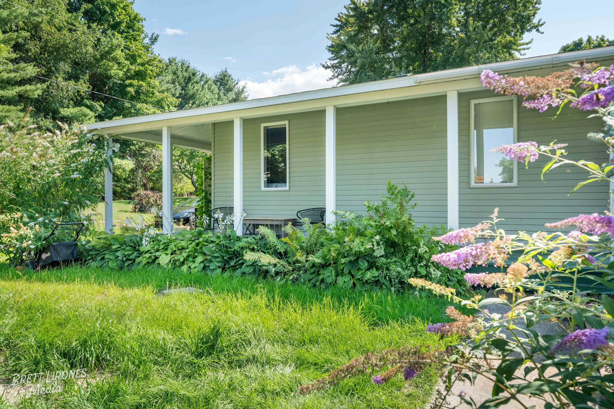 Side view of a house with a green exterior, white trim, and a covered porch, surrounded by green plants and trees on a sunny day.