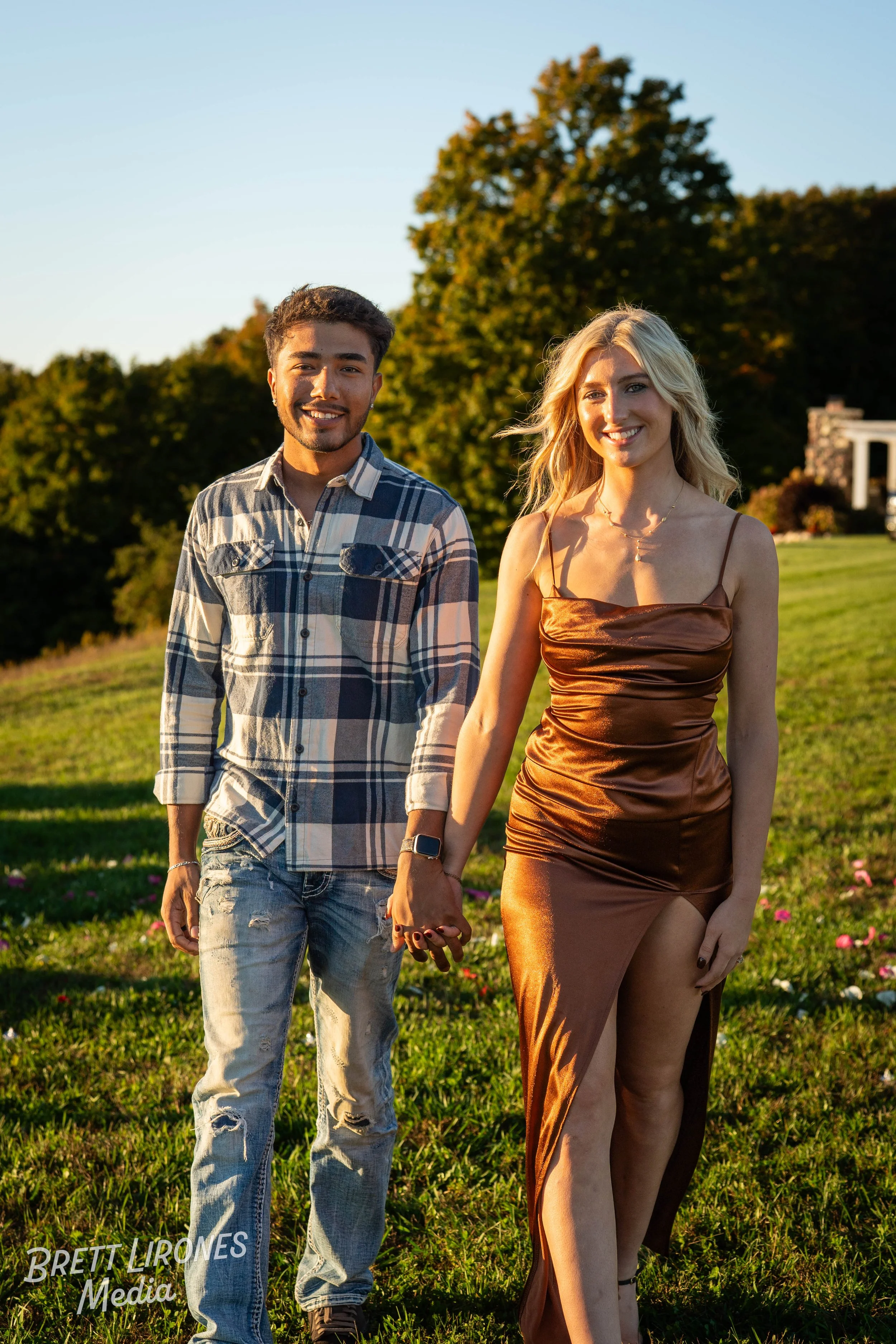 A smiling young couple holding hands and walking outdoors in a grassy field during sunset, with trees in the background.