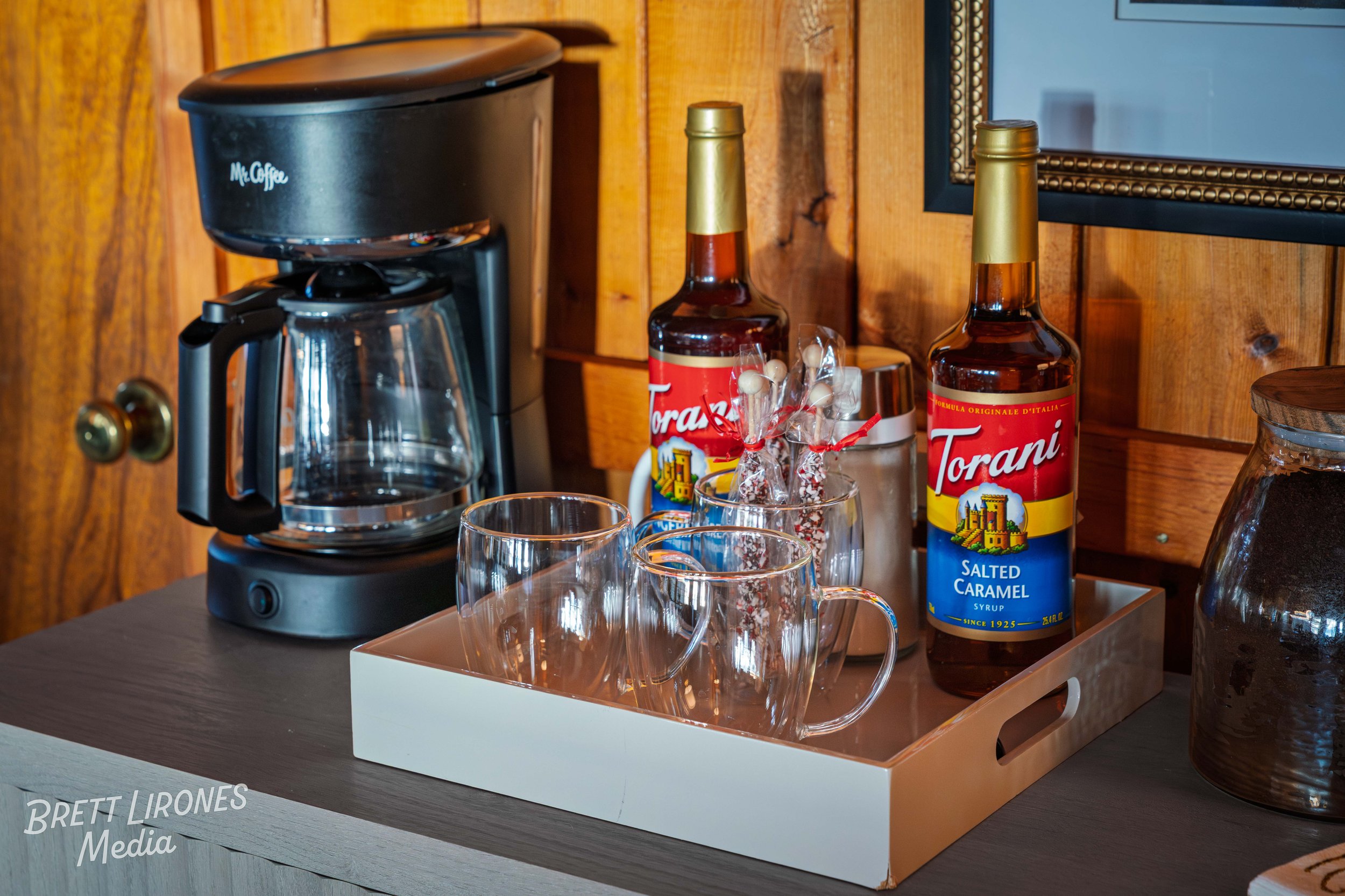 A coffee station on a wooden counter with a Mr. Coffee drip coffee maker, two small bottled syrups labeled Torani Salted Caramel, three clear glass mugs, and a small container of assorted candies in a glass jar.