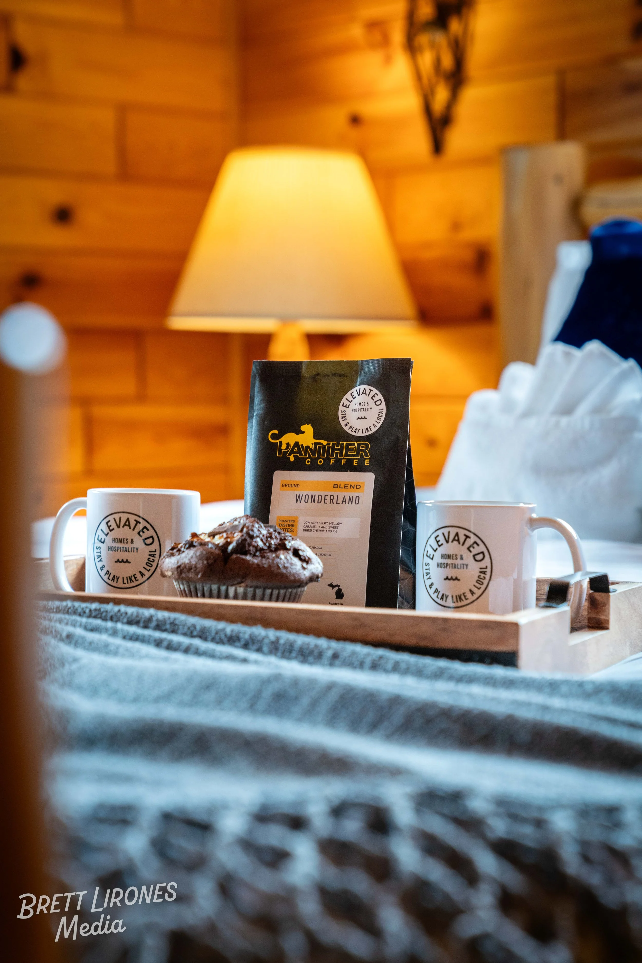 Tray with two white mugs bearing a circular logo, a muffin dusted with powdered sugar, and a bag of Panther Coffee blend, set on a bed with a safety clip attached, against a cozy wooden-paneled room with a warm lamp in the background.
