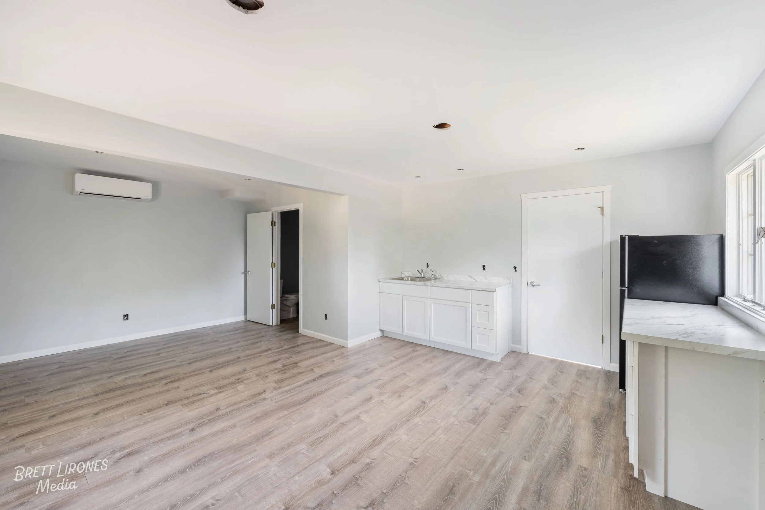 Empty room with light wood flooring, white walls, and a ceiling with recessed lighting. There is a white cabinet with a marble countertop and a sink. A black refrigerator is next to a window, and there is an open door leading to a bathroom with a vis