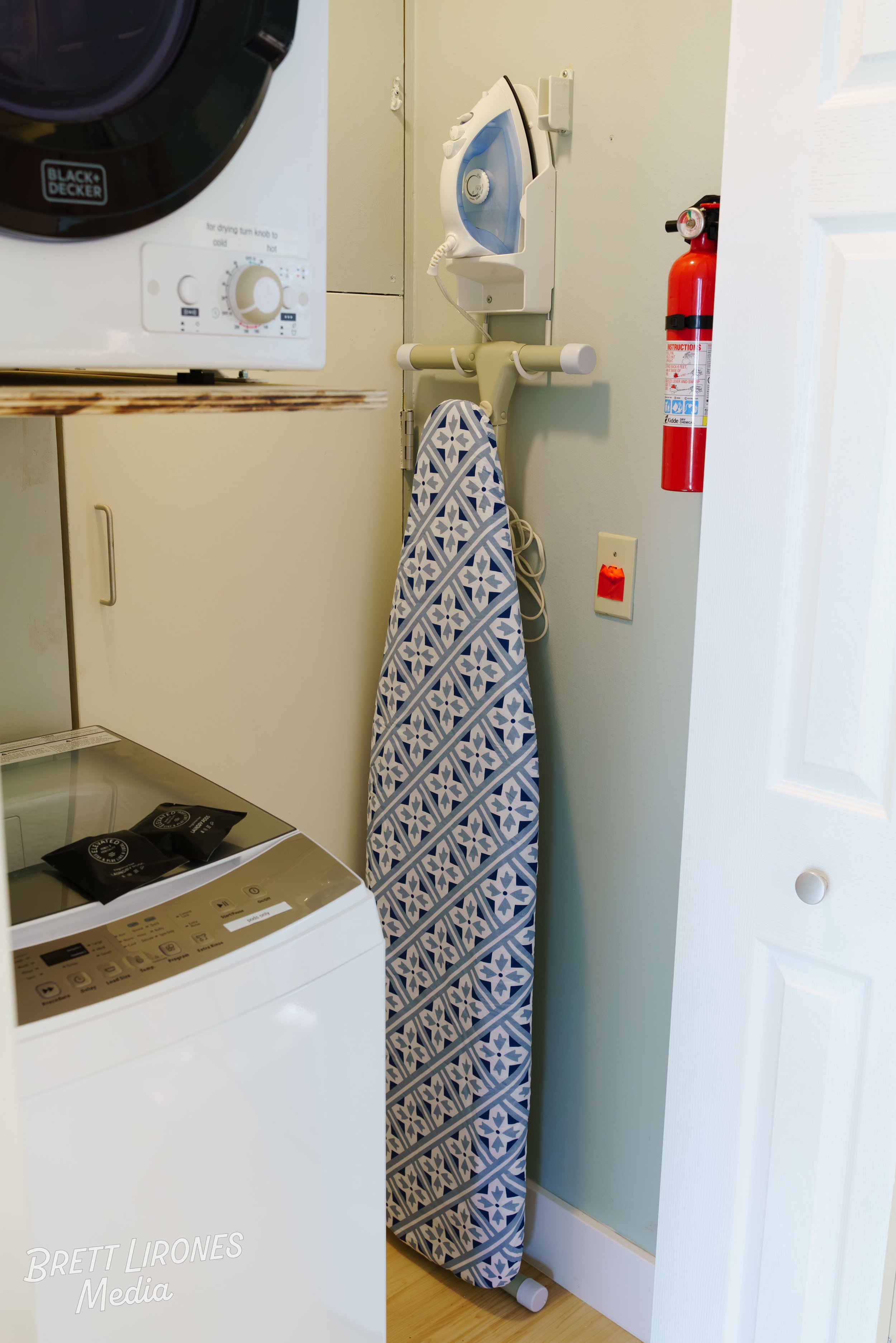A laundry room with a black Black & Decker dryer, a white wall-mounted steam iron, a blue and white patterned ironing board, a fire extinguisher, and a washing machine with two black packets on top.