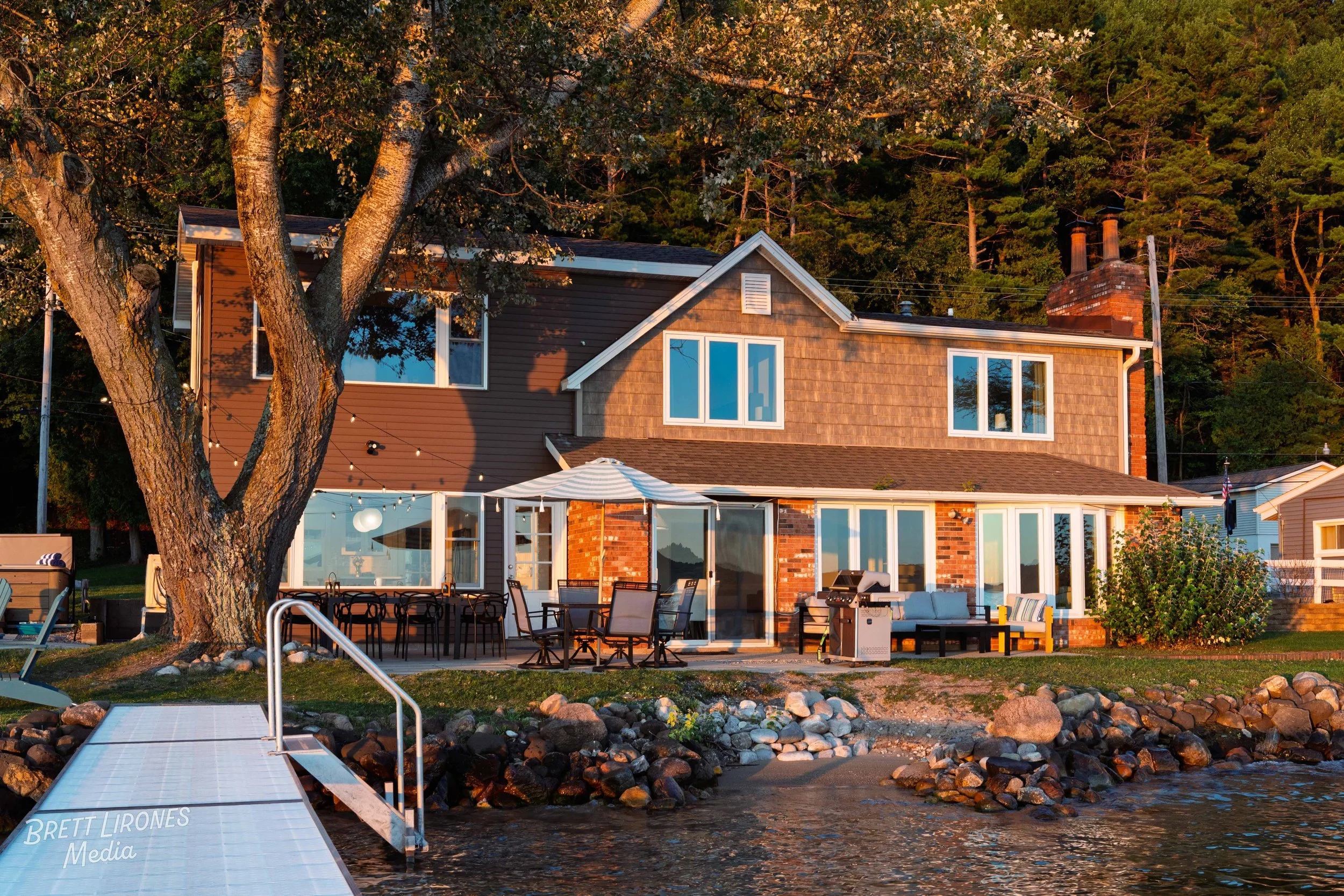 A two-story house by the water with a boat dock, surrounding trees, outdoor patio furniture, umbrella, and string lights, during sunset.