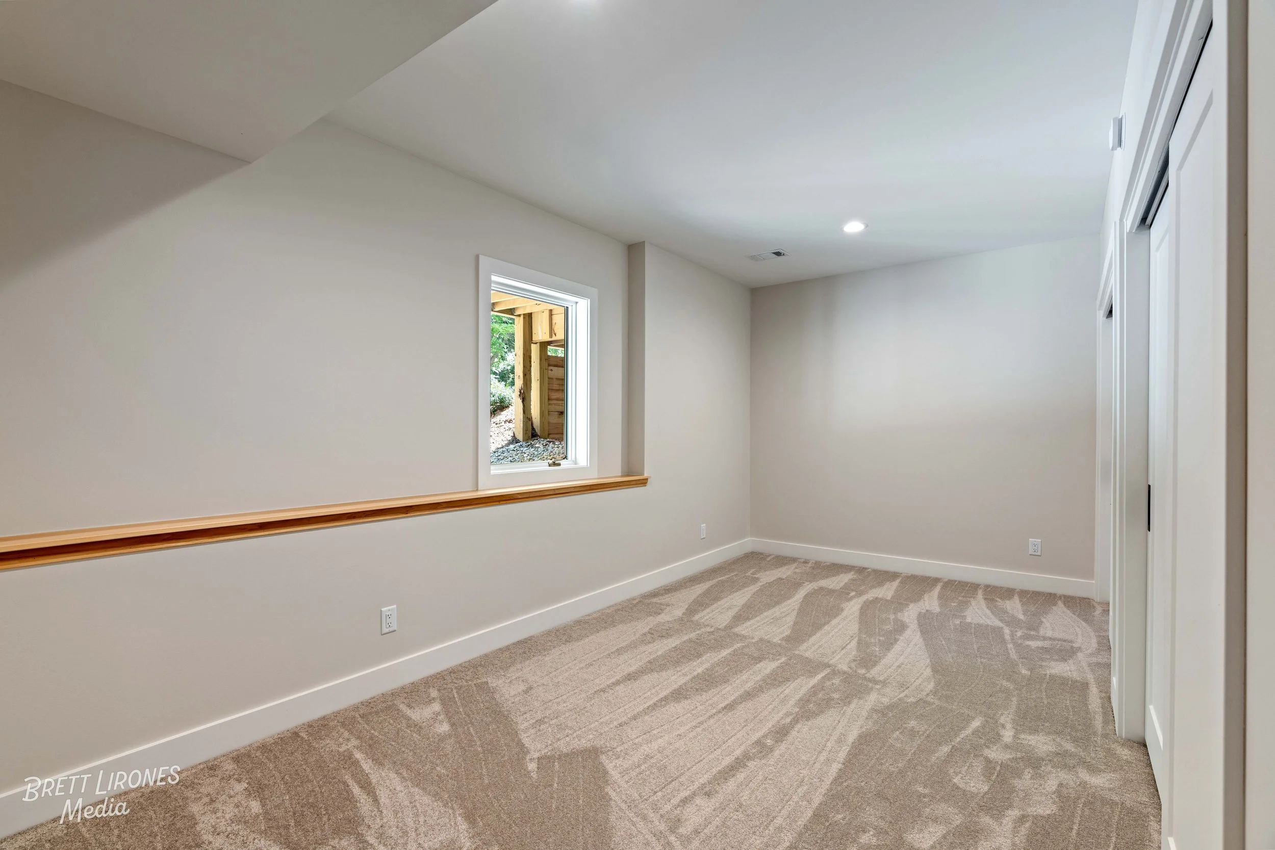 Empty beige carpeted room with a small window showing a wooden fence outside, plain white walls, ceiling-mounted lights, and closet doors.