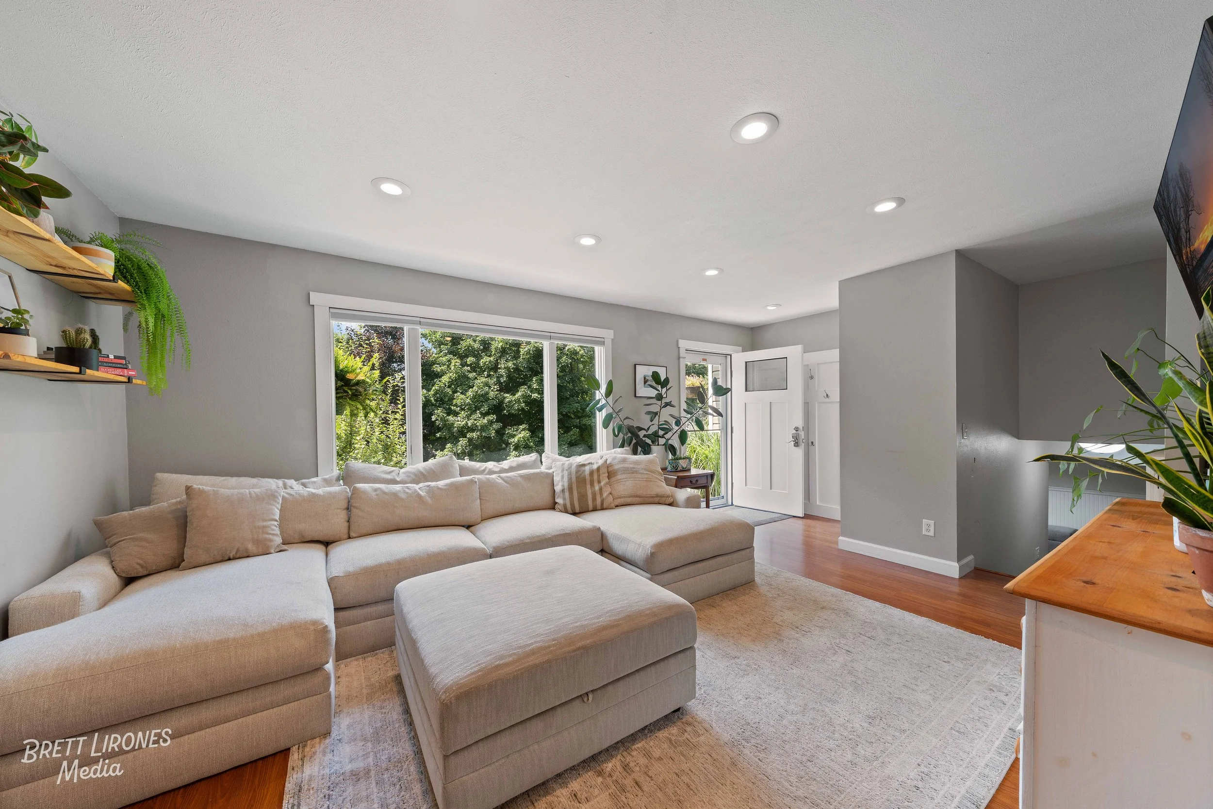 Living room with large beige sectional sofa, a beige ottoman, potted plants near window, wooden shelves with plants and books, hardwood floor, white walls, and front door in background.