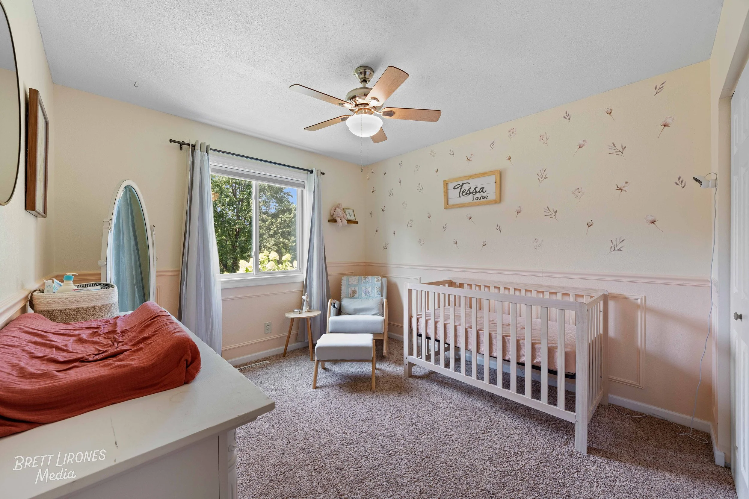 Nursery room with a crib, armchair, and changing table, decorated with floral details and a window with curtains.