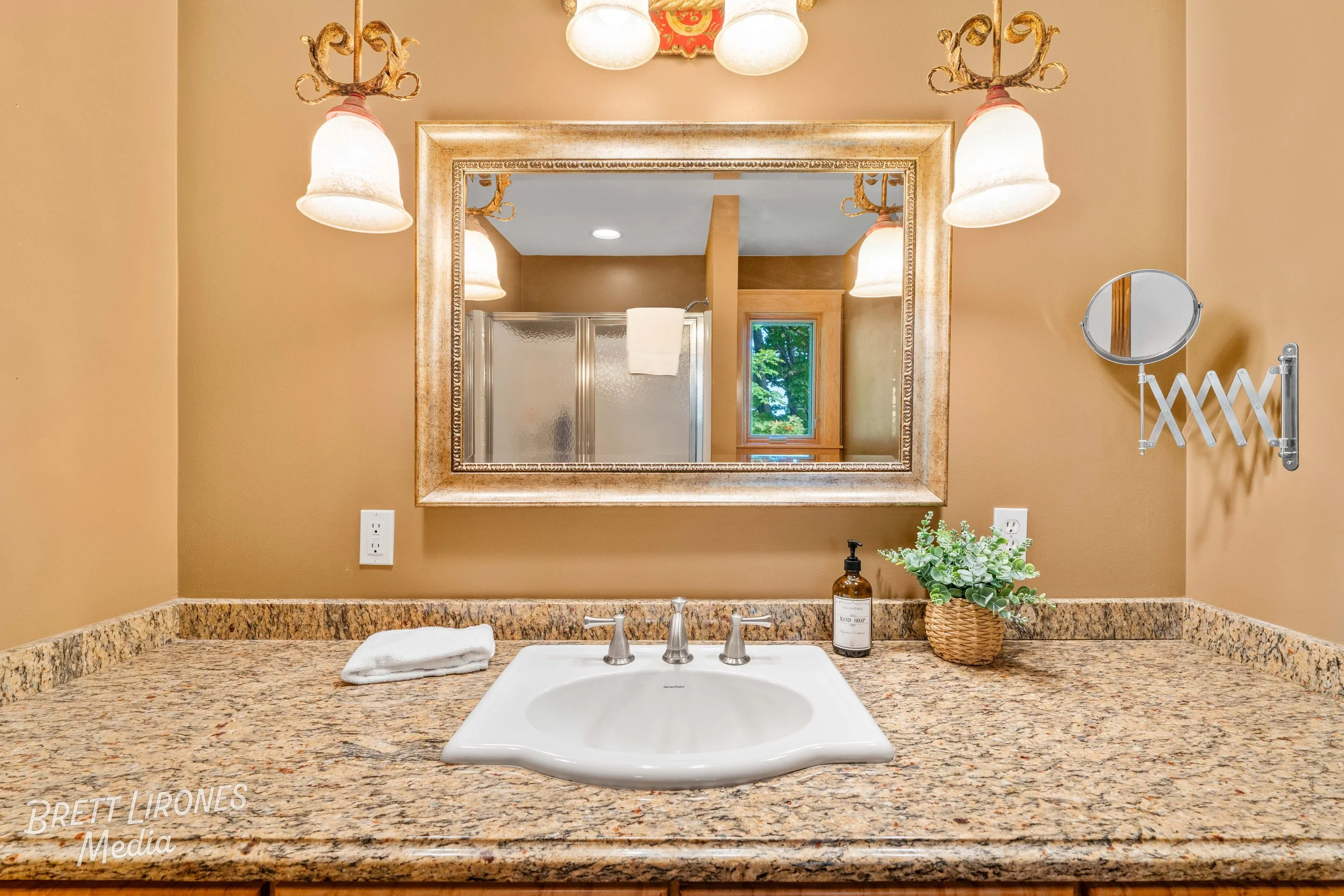 Bathroom vanity with a granite countertop, a white sink, a mirror, wall-mounted light fixtures, a small mirror on an adjustable arm, a bottle of hand soap, a small potted plant, a towel, and a bathroom visible in the reflection.