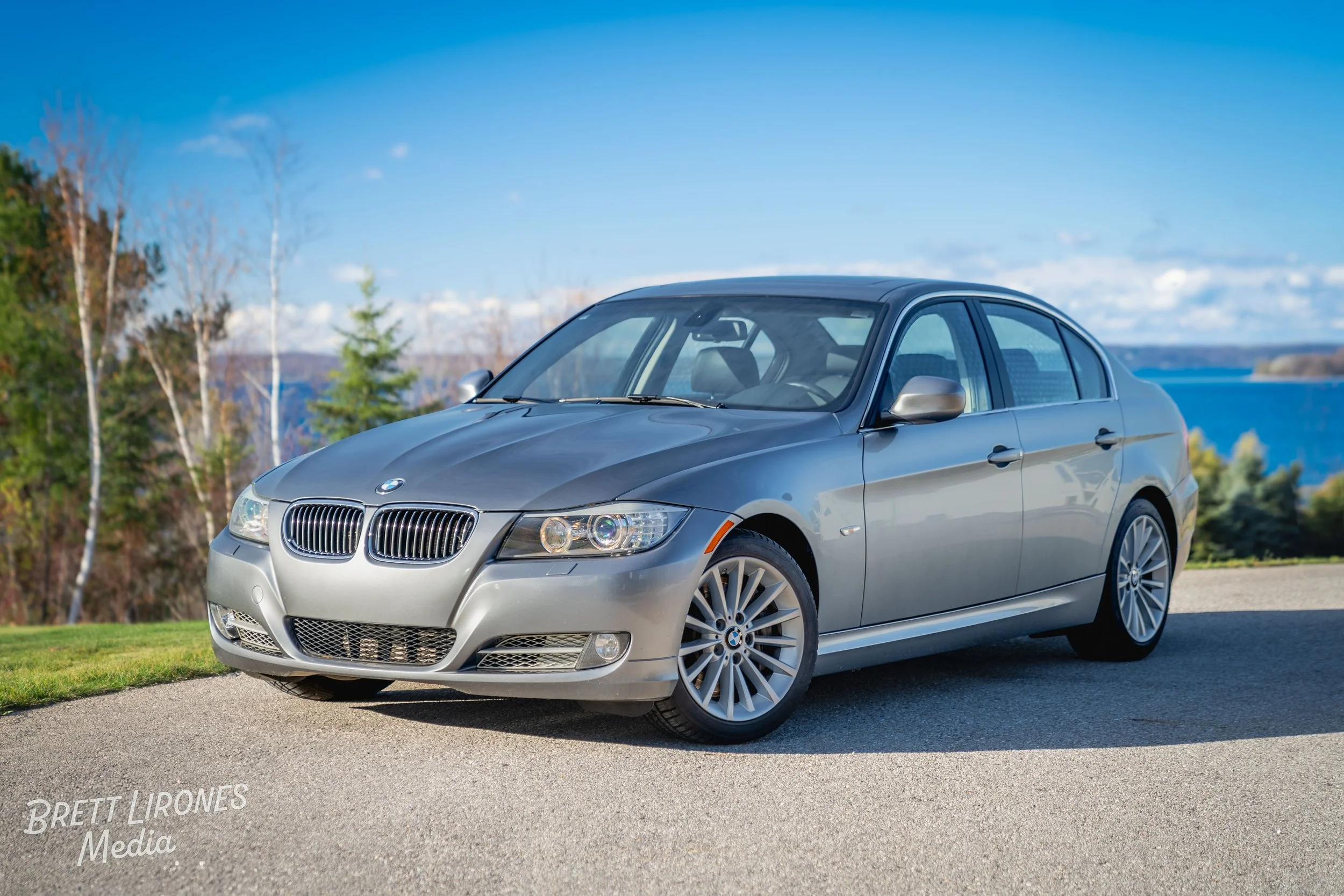 Silver BMW sedan parked on the side of the road with trees, a lake, and a partly cloudy sky in the background.