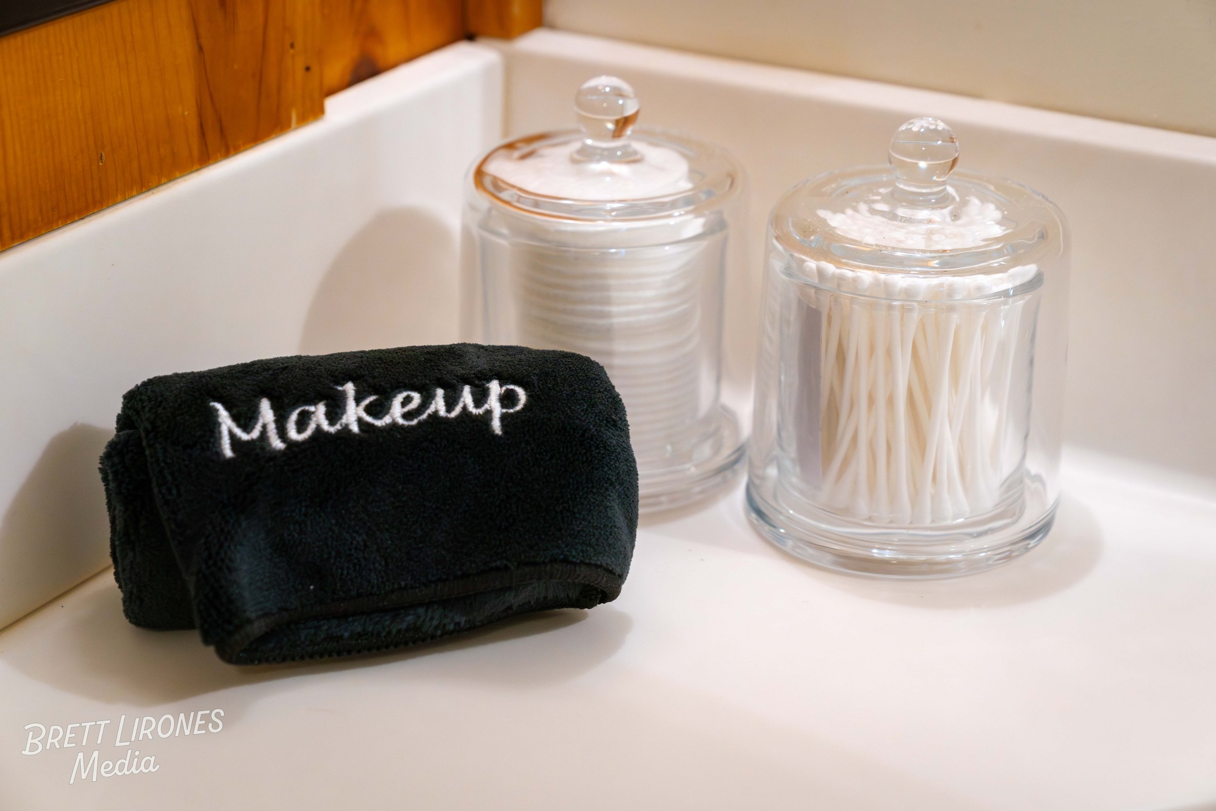 A makeup remover cloth with the word 'Makeup' embroidered on it, two glass containers with glass lids, one holding cotton pads and the other holding cotton swabs, all placed on a white surface near a wooden wall.