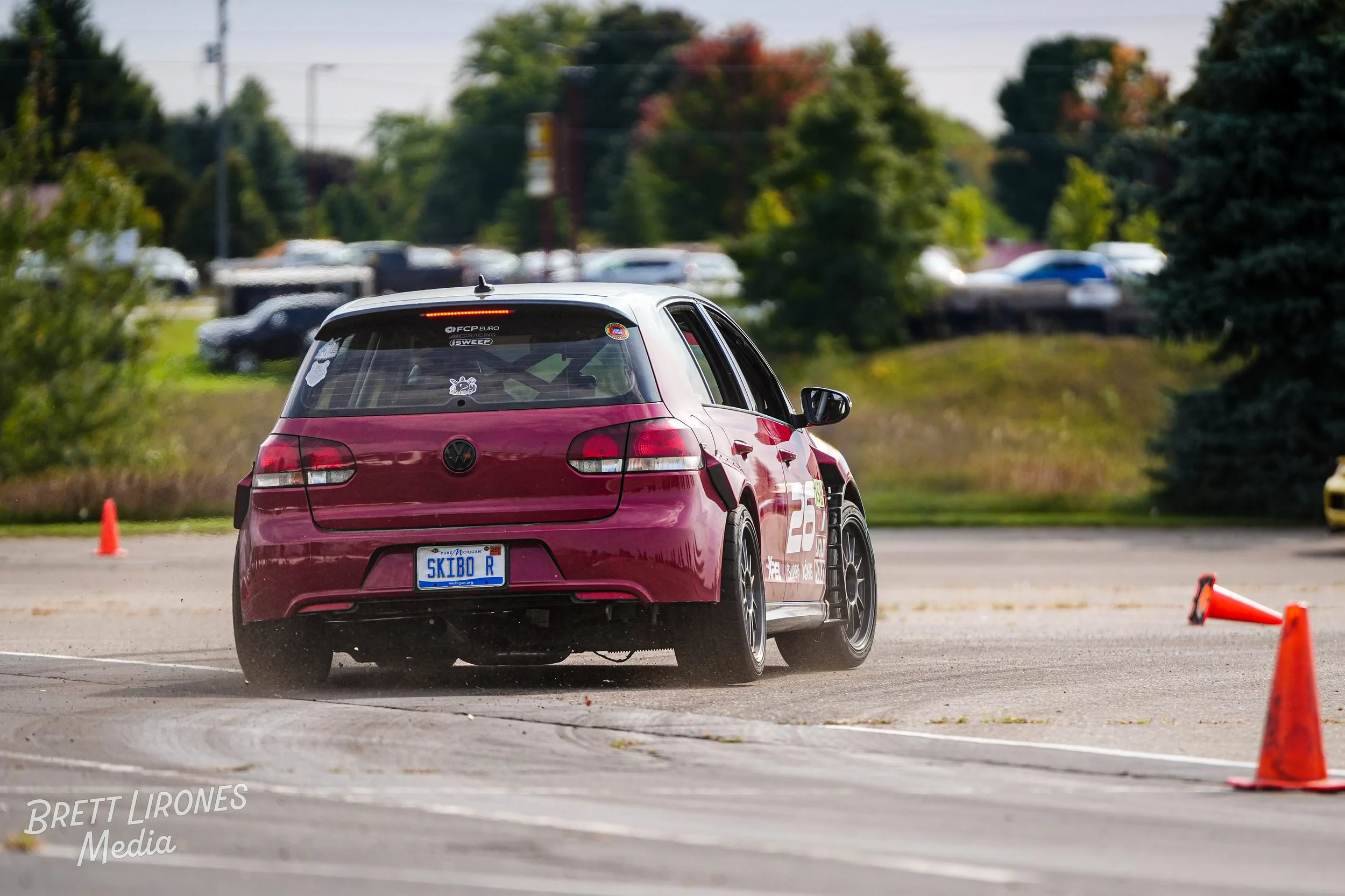 Red Volkswagen car navigating a cone course in an open parking lot during a driving event, with other cars and trees in the background.