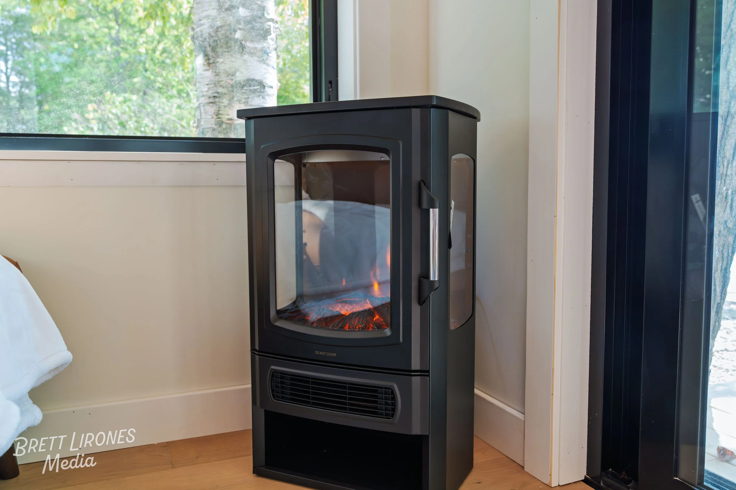 A black electric fireplace heater in a room with wooden flooring, near a window showing green trees outside and a glass door on the right.
