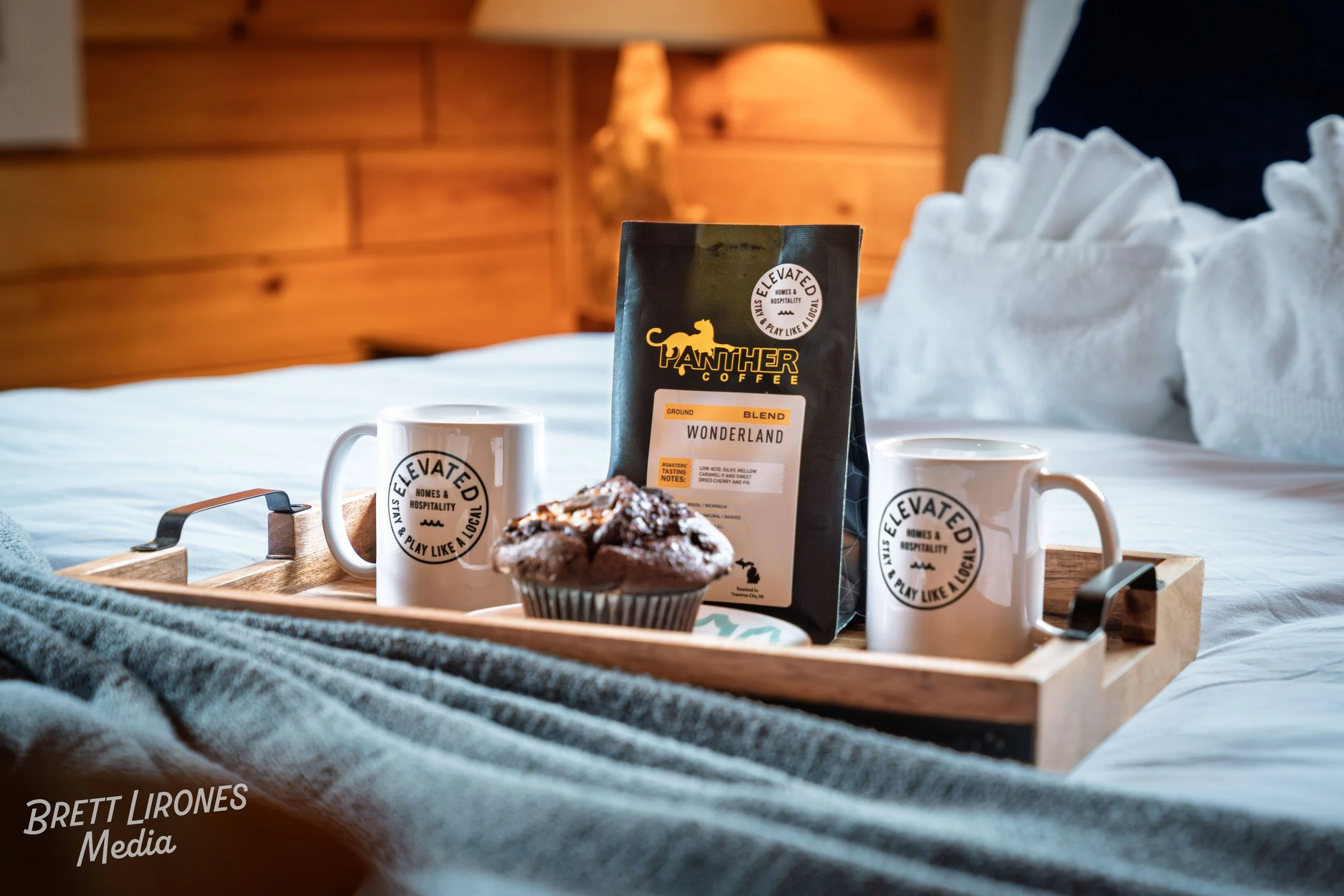 Travel tray with two white mugs labeled 'ELEVATED HOMES & HOSPITALITY' holding coffee, a chocolate muffin, and a bag of Panther Coffee Wonderland blend, all on a bed with a gray blanket in a cozy wooden room.