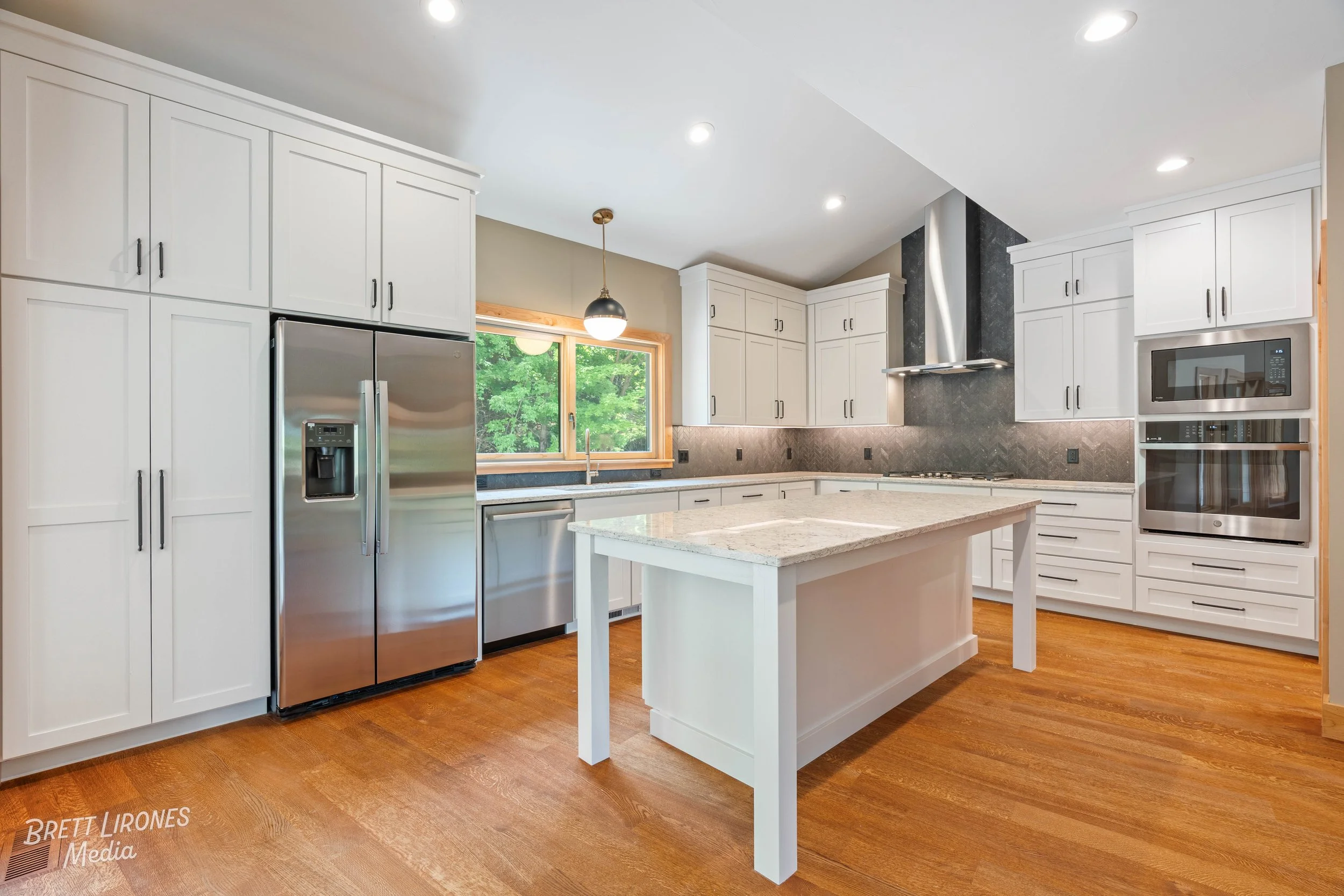 Modern kitchen with white cabinets, stainless steel appliances, a kitchen island, wooden flooring, a window showing green trees, and recessed lighting.