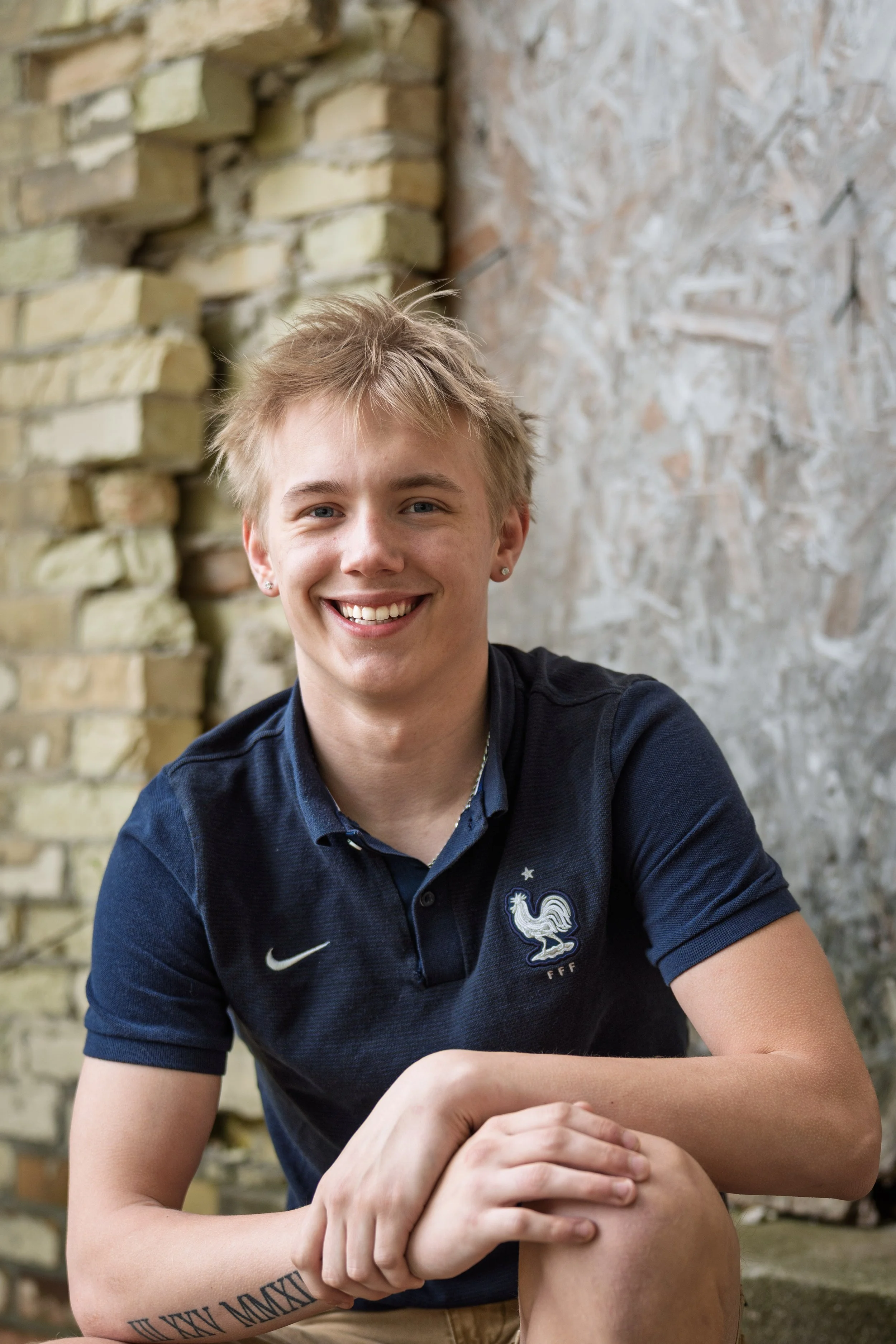 A young man with blond hair smiling at the camera. He is wearing a navy blue polo shirt with a logo of a rooster and the French Football Federation emblem. He is sitting outdoors against a background of a stone wall and textured concrete wall.