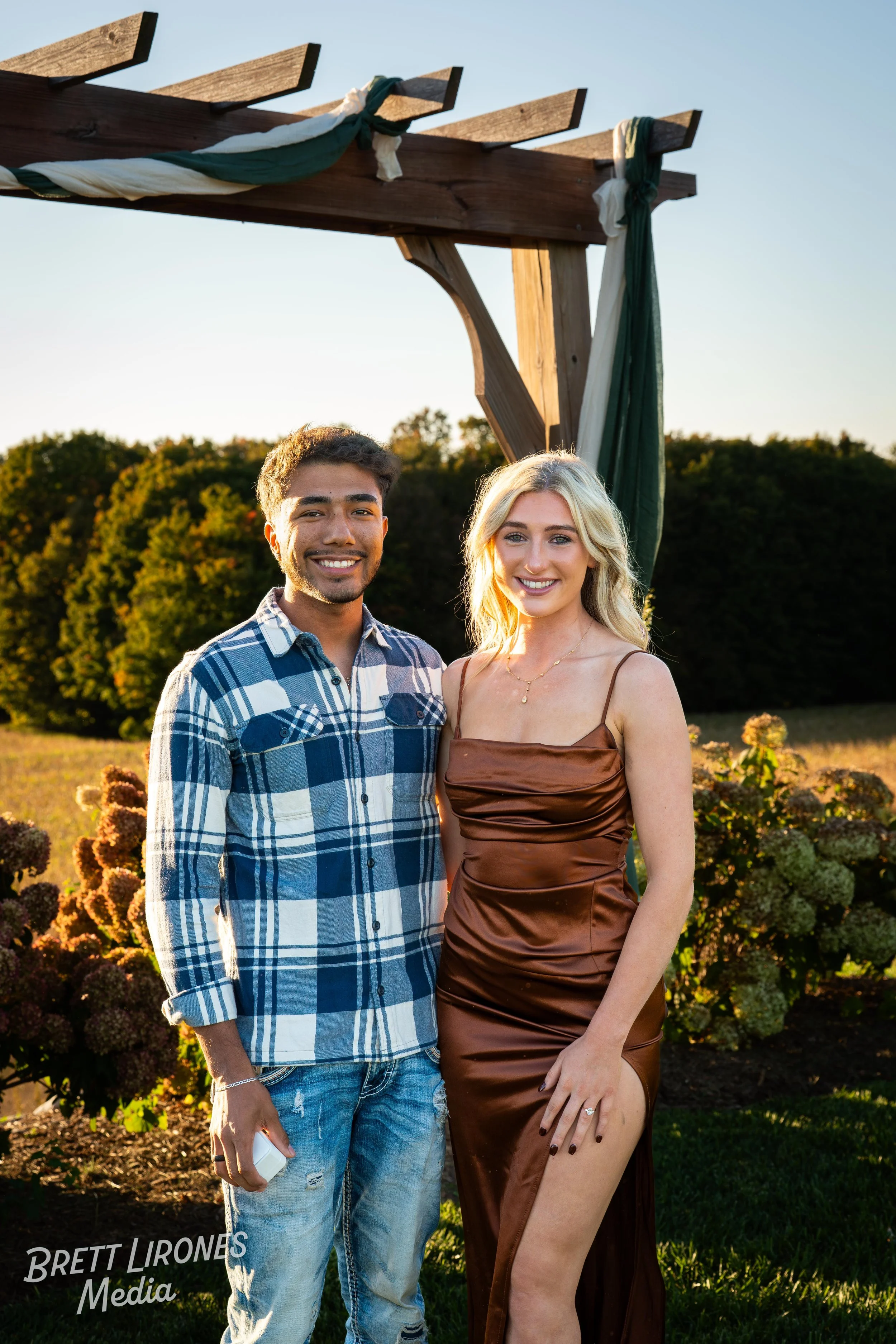 A young man and woman posing outdoors, smiling, in front of a wooden arch with fabric decorations, with trees and a field in the background, during sunset.