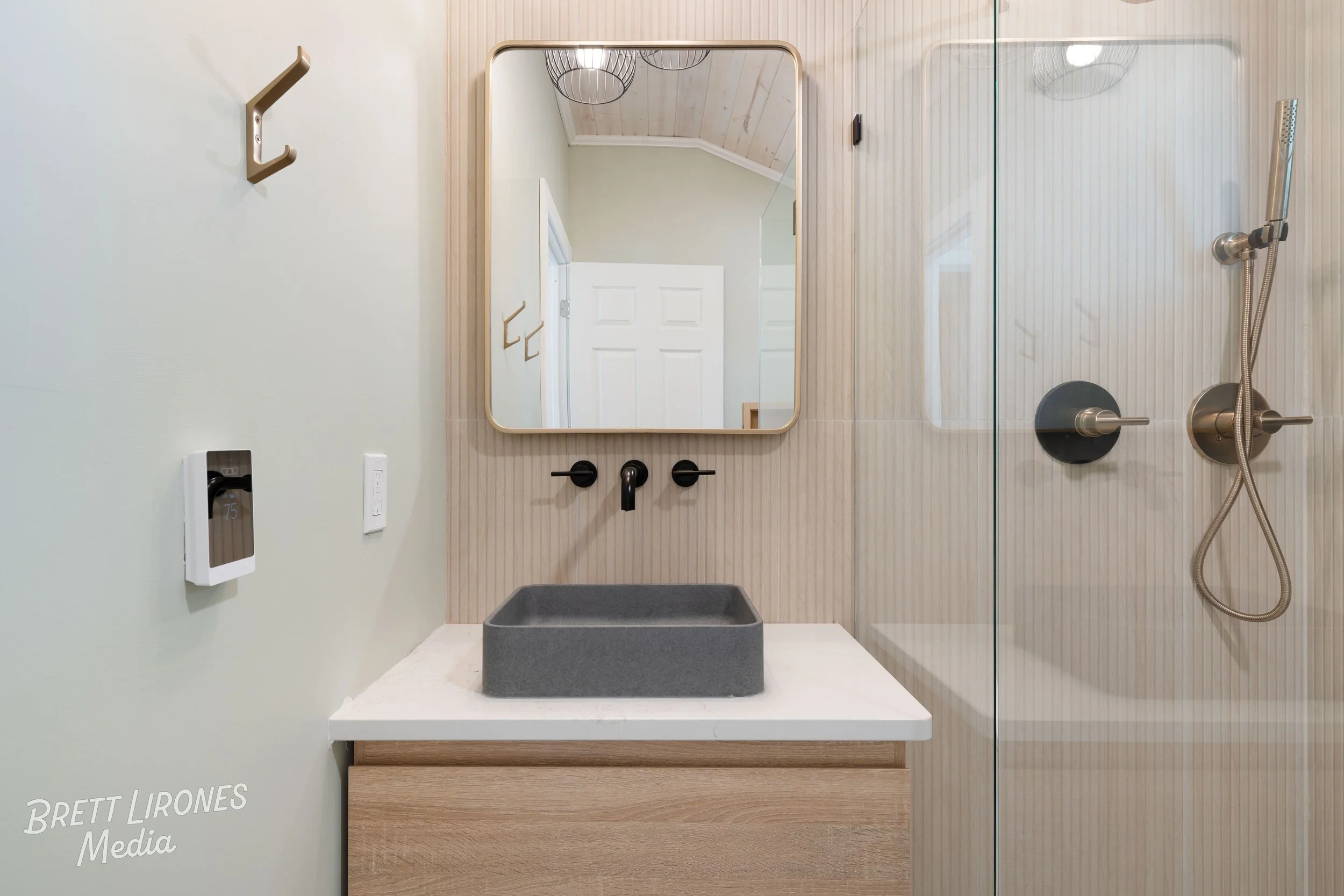 Modern bathroom with a mirror, a gray vessel sink on a white marble countertop, black fixtures, a walk-in shower with a handheld showerhead, and a white wall with wooden accents.